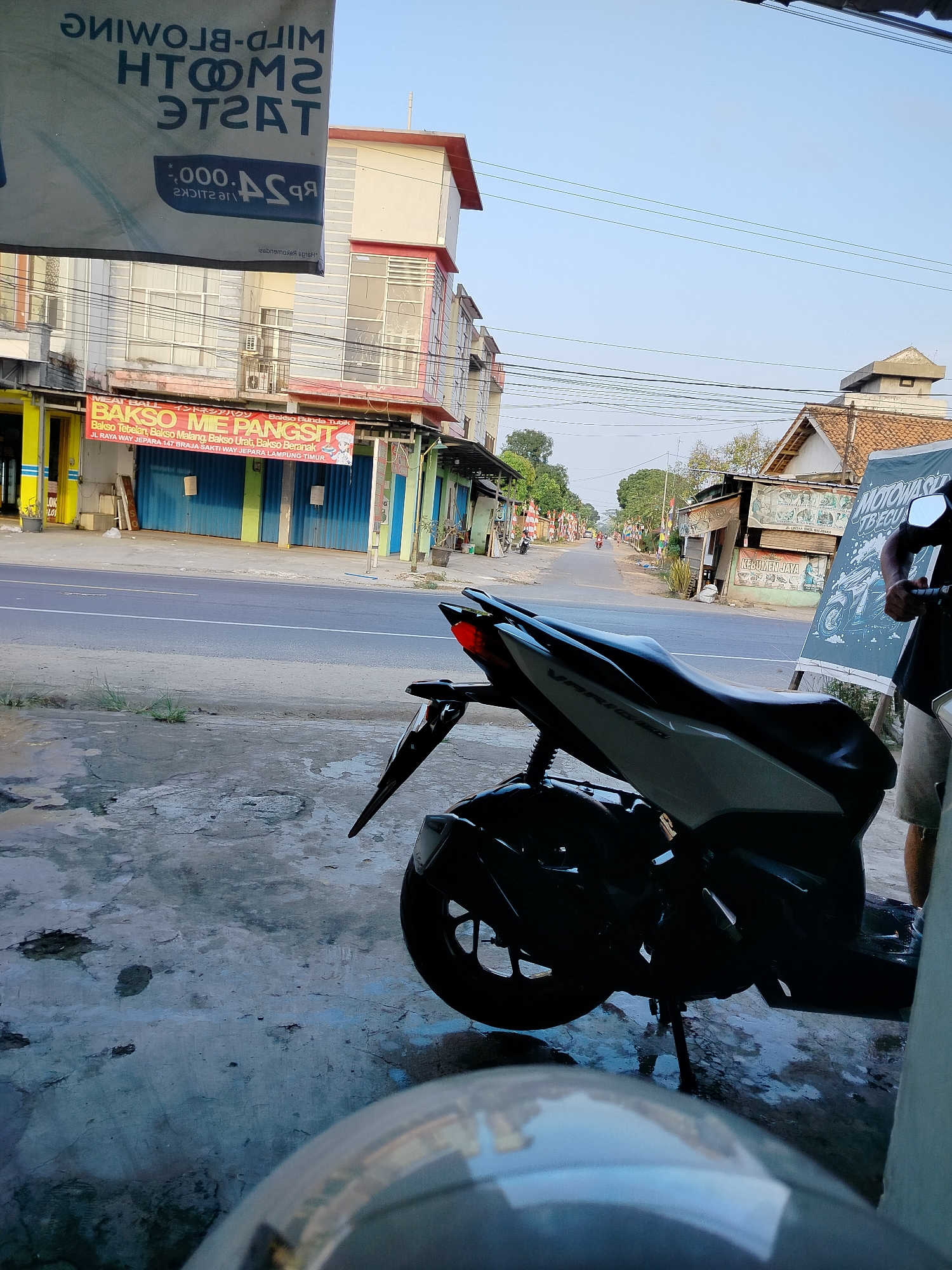 The image depicts a street scene in Braja Asri, Indonesia. In the foreground, a black and white scooter is parked on a concrete surface with puddles of water. The scooter's tail light is visible, and its license plate is obscured. To the right, a portion of a person wearing light-colored shorts is visible, holding onto the scooter's handlebars. Behind the scooter, a street stretches into the distance, lined with buildings. One building has a prominent banner that reads "BAKSO ME PANGSIT" and lists various types of meatballs and their prices, along with an address on "JL. RAYA WAY JEPARA 147 BRAJA SAKTI WAY JEPARA LAMPUNG TIMUR." Further down the street, there are more buildings, some with awnings and storefronts, and a few people are visible, along with motorcycles and flags. The sky is clear and blue, indicating daytime. The overall impression is of a commercial street in a moderately populated area.