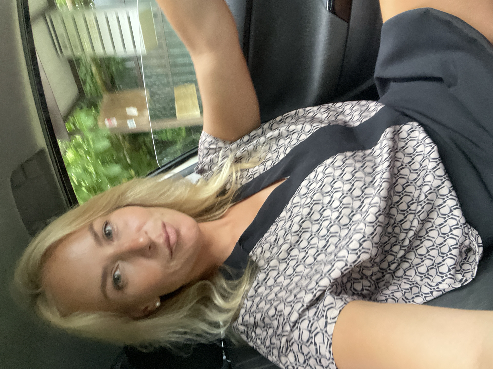 A blonde woman is pictured inside a car, looking forward and to the left. She is wearing a patterned top with a black neckline and a black skirt. The woman has blonde hair and is wearing a pearl earring in her left ear. The interior of the car is visible, including the dashboard and the side door. Outside the car window, trees and structures are visible, suggesting an outdoor location, possibly in a suburban or rural area. The overall scene captures a moment of the woman driving or sitting in the passenger seat.