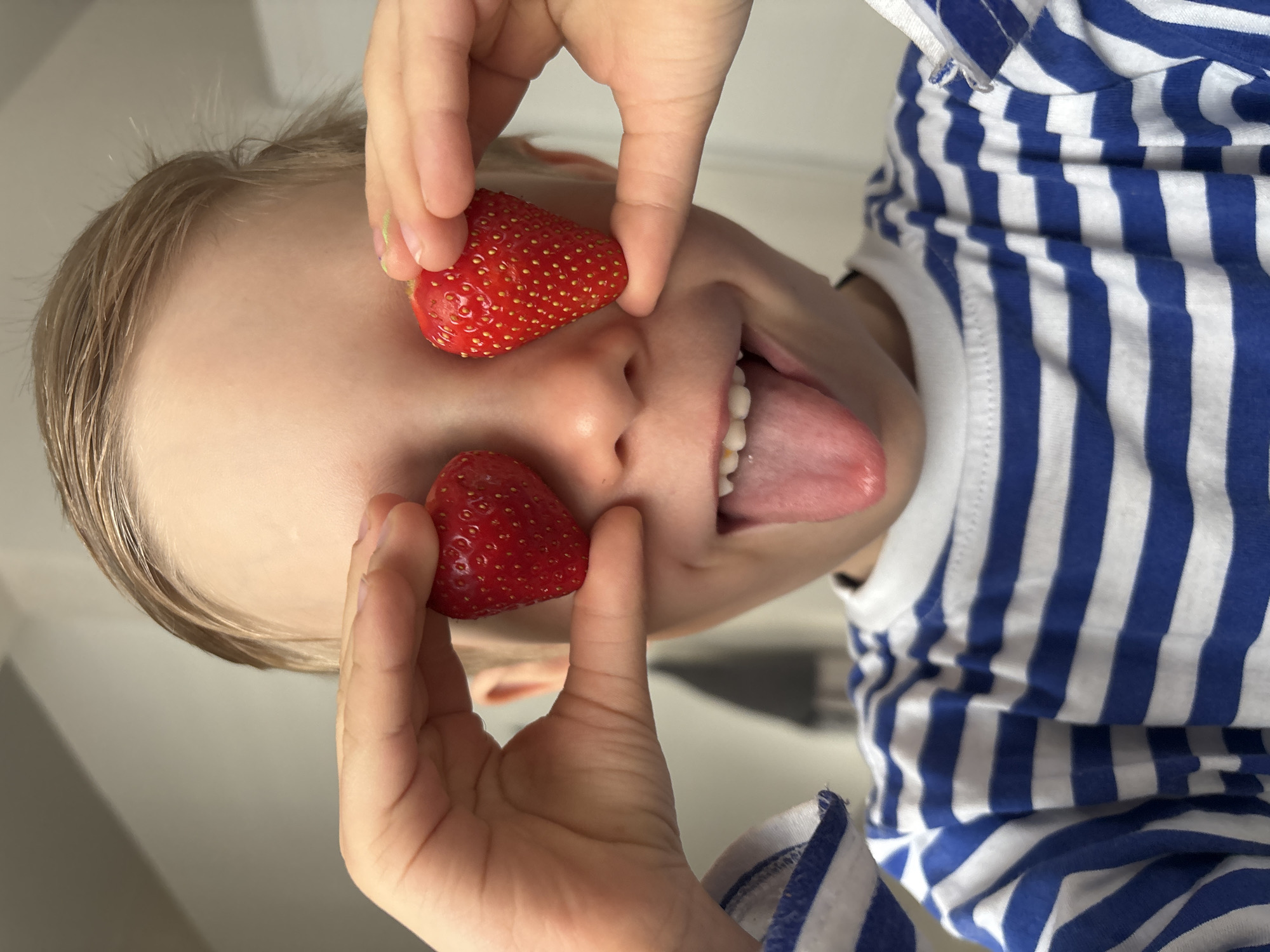 A child with blond hair is upside-down, sticking out their tongue, and someone is holding two strawberries up to their eyes as if they are glasses. The child is wearing a blue and white striped shirt. The location is unknown.