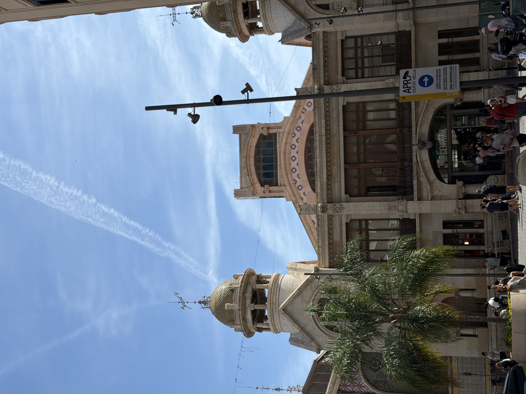 This image captures a vibrant street scene dominated by the Mercat Central (Central Market) in Valencia, Spain, under a clear blue sky. The market building, a prominent example of Valencian Art Nouveau architecture, features a light-colored stone facade with intricate detailing. Two distinctive golden-domed towers, topped with decorative weathervanes, frame the structure, with one fully visible on the left and another partially on the far right. The central section of the building has a lighter, possibly tiled roof decorated with a pattern of small blue and white circular motifs. Large arched windows are visible on the upper levels, with wooden frames.
In the lower center, the main entrance to the market is an arched doorway with "Mercat Central" inscribed above it. A crowd of people is either entering or exiting the building. Numerous individuals are scattered across the street and sidewalk in the foreground and midground, dressed in casual summer clothing. They appear to be walking, standing in small groups, or congregating near the market entrance.
A large, multi-trunked palm tree with extensive green fronds stands in the lower-left quadrant, partially obscuring the building's facade. A tall, dark grey lamp post with multiple downward-facing light fixtures is prominent in the right-center foreground. Attached to this pole is a rectangular yellow and blue sign indicating "APR CIUTAT VELLA" (Residential Priority Area - Old City), "EXCEPTE AUTORITZATS / EXCEPTO AUTORIZADOS," and "ZONA VIDEOVIGILADA," signifying a restricted access area and surveillance.
The sky is bright blue with scattered, wispy white clouds. Two distinct airplane contrails diagonally cross the upper sky, originating from the upper left and extending towards the upper right. The scene is brightly lit by sunlight, indicating a clear, sunny day. Partial views of vehicles, including a dark car on the far left and a scooter on the far right, are also visible.