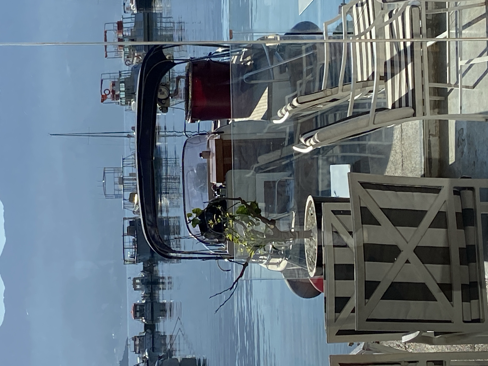 A reflected view through a window shows a water scene with various boats and a distant, faded coastline, likely captured in Budva, Montenegro. The reflection includes interior elements like striped patio chairs, a plant in a clear vase, and part of a boat or structure's framework. The reflections superimpose the distant waterfront scene onto the interior space.