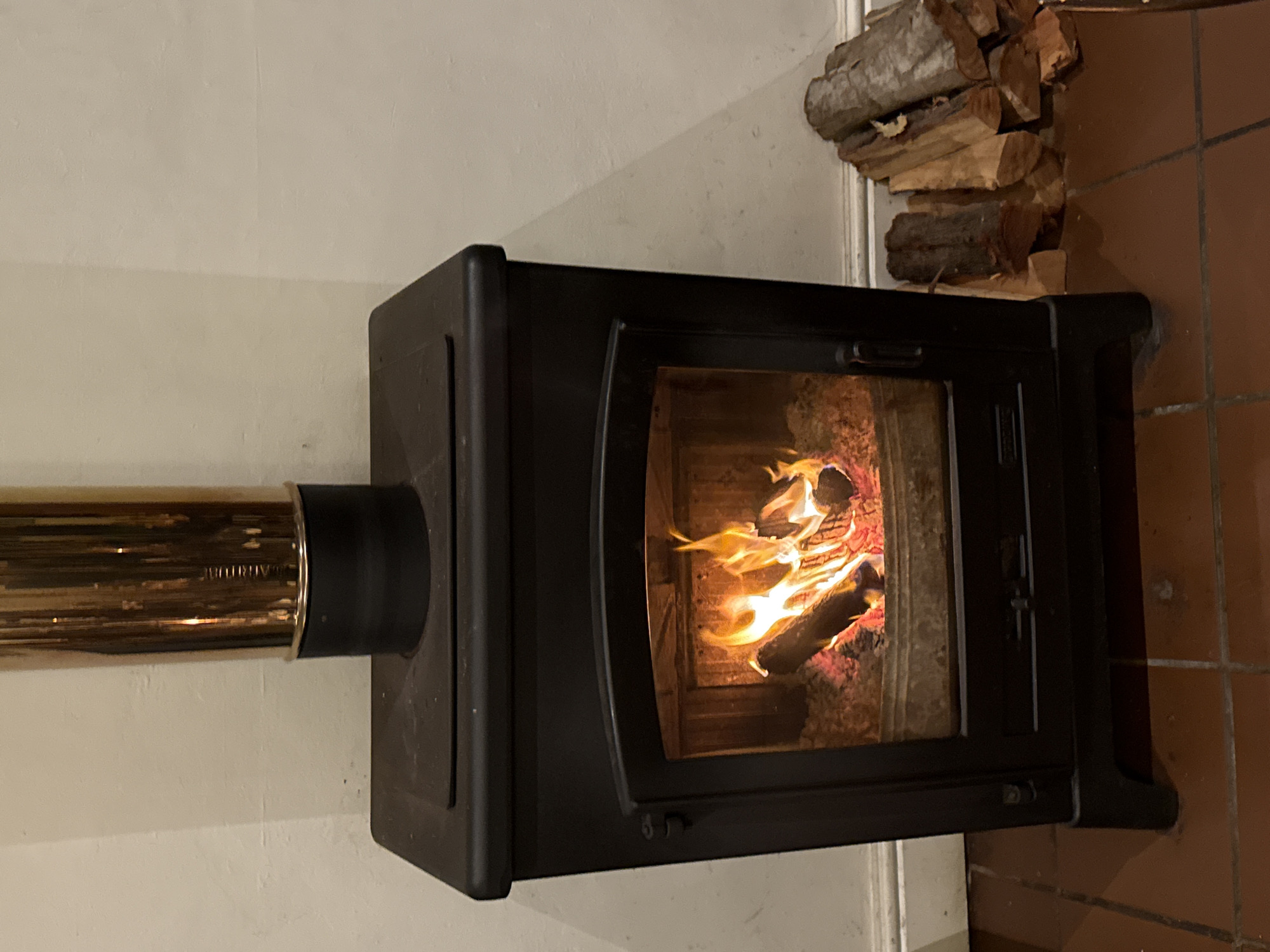 A slightly tilted medium shot features a black wood-burning stove with a fire burning inside. A golden metal pipe extends from the stove's left side toward the wall. To the upper right of the stove, a stack of firewood rests on the floor. The floor itself is tiled in terracotta. The walls are a light, neutral color. There is no indication or evidence that it is located in Paarl, South Africa.