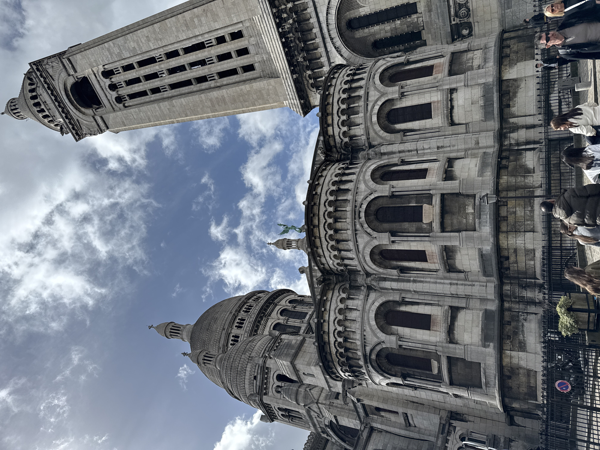 The image shows the Sacré-Cœur Basilica in Paris, France, taken from a low-angle perspective. The basilica's dome and bell tower are visible against a sky with white clouds. The building's exterior is made of light-colored stone with many arches and windows. A few people are seen in the foreground, near a black metal fence. The photograph appears to be taken during daylight.