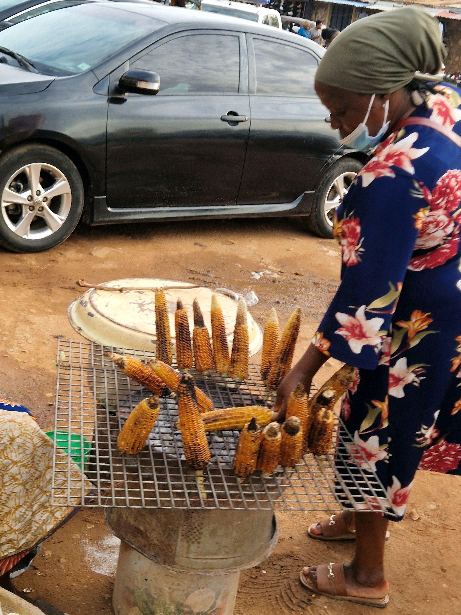 In Unknown, Unknown, a person wearing a green headscarf and a floral print dress is tending to corn being grilled over a metal grate. The corn appears to be roasted and is arranged on the grate, with some pieces showing char marks. A black car is parked behind the grilling setup, with its wheel and side visible. The ground is dirt, and there are various objects scattered around, including a metal tray and a green bucket. The person is also wearing a face mask, which is partially pulled down. The scene suggests an outdoor food vending operation.