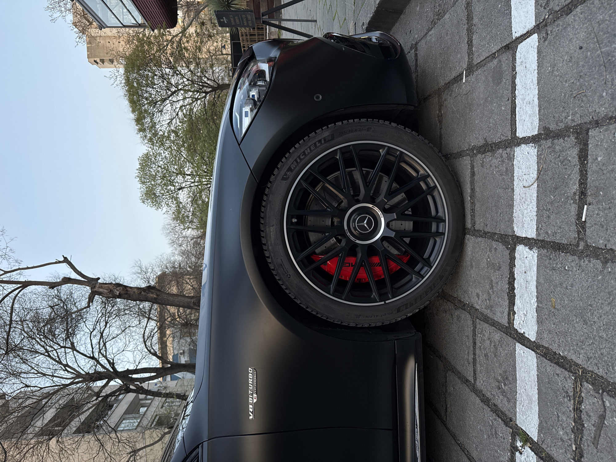 The image shows a close-up of the front of a matte black Mercedes-AMG car, specifically the front left wheel and part of the body. The tire is a Michelin Pilot Alpin. The wheel has a black multi-spoke design with red brake calipers visible. The "V8 BITURBO 4MATIC+ PERFORMANCE" emblem is on the side panel. The car is parked on a paved surface with a white parking line. In the background, there are buildings and trees, including an old stone tower. Based on the tower and architectural style, the location is likely Plovdiv, Bulgaria, specifically near the old town area. There are no people visible in the image. The focus is on the car's details.