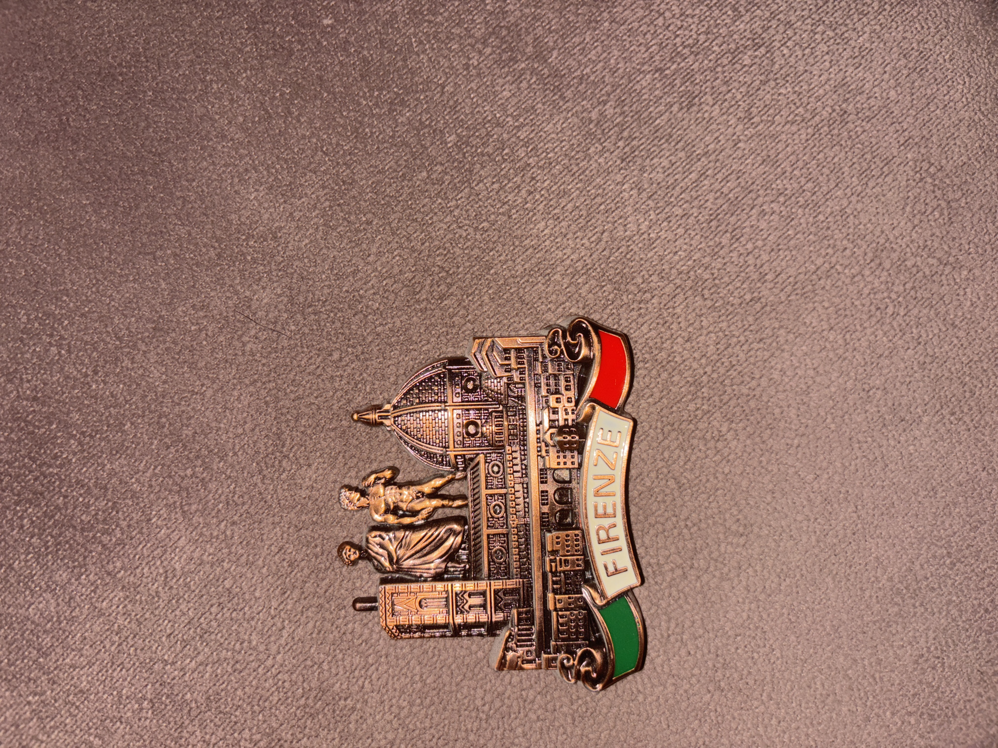 A close-up photograph shows a souvenir magnet depicting the city of Florence ("Firenze" in Italian). The magnet, made of a bronze-colored metal, features representations of the Duomo (Florence Cathedral) with its iconic dome, and sculptures. It also includes a banner with the name "FIRENZE" written on it, flanked by the colors of the Italian flag: red and green. The magnet sits on a surface with a textured, brown background. There are no people or any indications of being in Montana, Bulgaria.