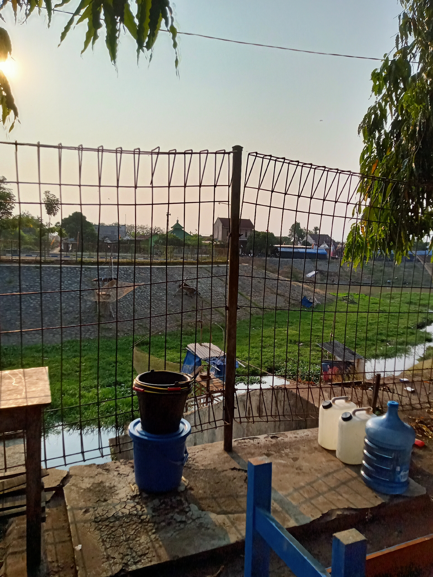 The image depicts an outdoor scene, likely in a suburban or rural area of Majan, Indonesia, as suggested by the presence of vegetation and modest structures. The foreground features a concrete area with two stacked buckets, one blue and one black, on the left. To the right, three large plastic containers are visible: two white jerrycans and a blue water dispenser bottle. A metal fence runs across the middle of the frame, separating the foreground from a grassy area beyond. In the distance, across the grassy area and a concrete embankment, several buildings, including a structure with a green roof resembling a mosque or religious building, are visible. Trees and foliage are present throughout the scene, with sunlight filtering through the leaves in the upper left corner, indicating it is likely late afternoon or early morning. The overall impression is a functional, possibly residential or community outdoor space, with a utilitarian focus on water storage.