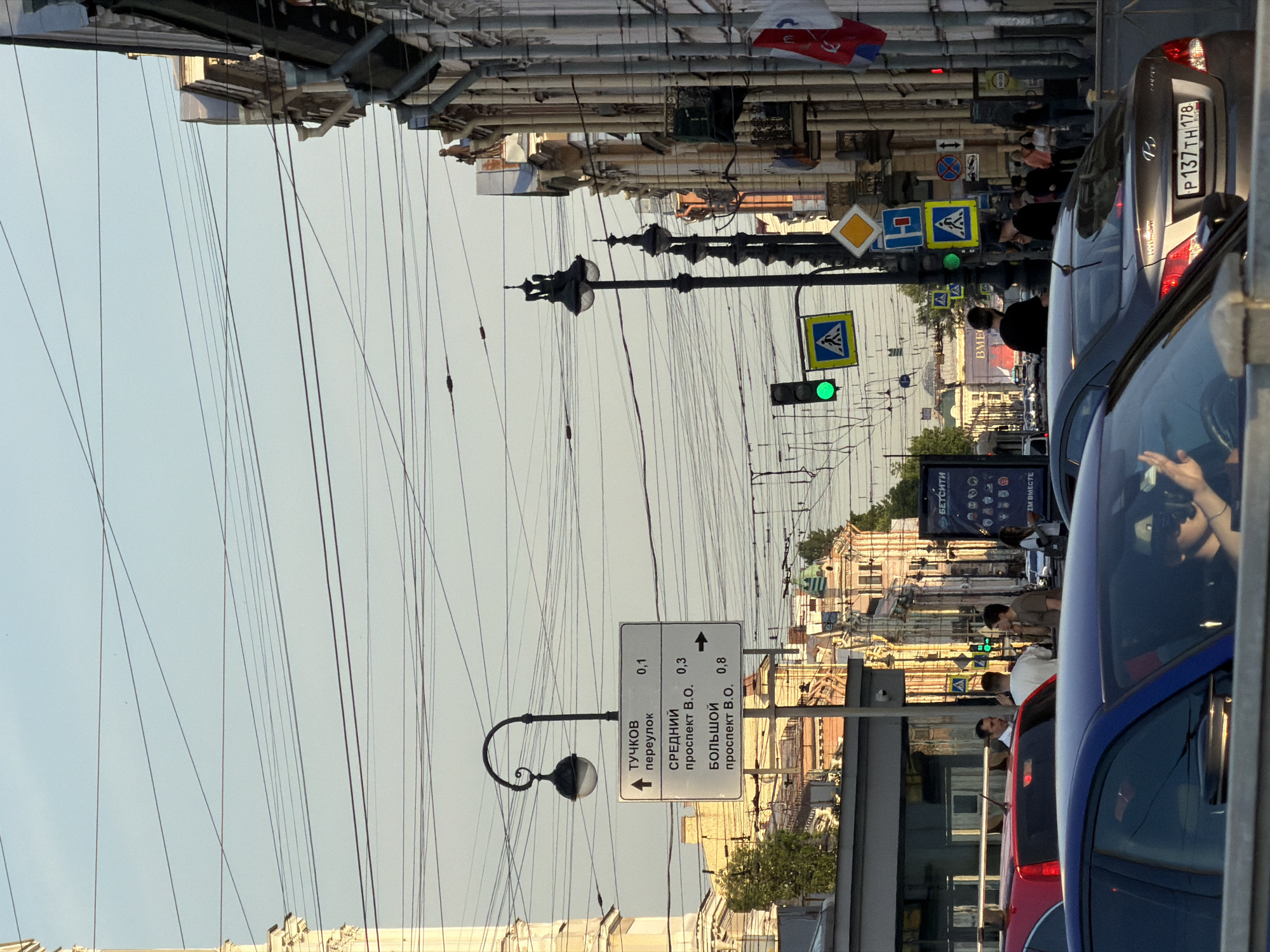 The image depicts a street scene in Saint Petersburg, Russia, likely near Palace Square or along Nevsky Prospekt, given the architectural style and the presence of street signs indicating directions to major streets such as Tuchkov pereulok, Sredny prospekt V.O., and Bolshoy prospekt V.O. (Vasilievsky Ostrov). The sky is clear and pale blue, suggesting it is either early morning or late afternoon.
The foreground is dominated by the hoods of several cars, with the license plate of a brown Hyundai sedan visible, reading "P 137TH 178". A person is visible driving this car, wearing sunglasses and holding what appears to be a phone. The middle ground features a busy street with multiple lanes of traffic. Pedestrians are walking on the sidewalk, and there are traffic lights and numerous street signs, including pedestrian crossing signs. A street lamp with a decorative fixture is prominent on the left side of the image, with a directional sign attached to its pole.
The background reveals the characteristic architecture of Saint Petersburg, with ornate buildings featuring classical facades, columns, and detailed ornamentation. Several flags are visible on the buildings, including what appears to be the Russian flag and another tricolor flag. The overall impression is one of an active urban environment with significant overhead wiring, typical of older city centers. The sunlight casts long shadows, indicating the direction of light.