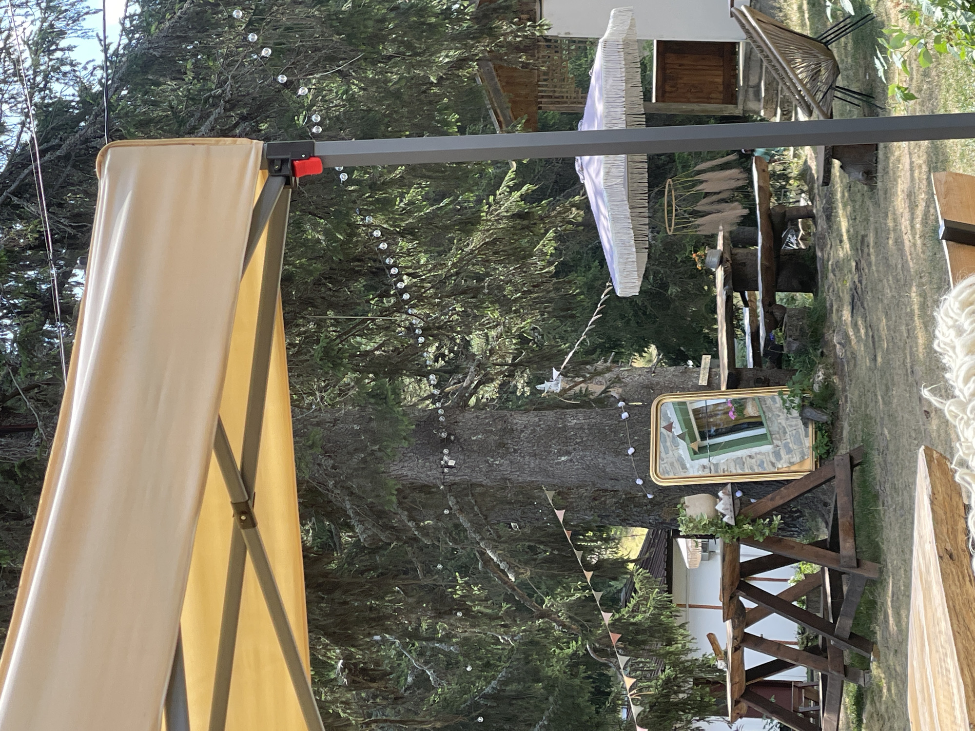 The image captures an outdoor seating area, possibly part of a cafe or guesthouse, located in the city of Smolyan, Bulgaria. A large tree dominates the center of the frame, draped with string lights and small triangular flags. To the left, a yellow canopy is partially visible, supported by metal poles, one of which has a red button. In front of the tree, a large, ornate gold-framed mirror reflects a stone building with a green-trimmed window. Wooden A-frame sawhorses are placed to the left of the mirror. Further into the scene, a rustic wooden picnic table with benches sits under a fringed umbrella. To the right of the table, a rattan chair is positioned on a stone patio leading to a wooden door. The ground is covered in dry grass, suggesting a warm, possibly summer day. No people are visible in the frame.