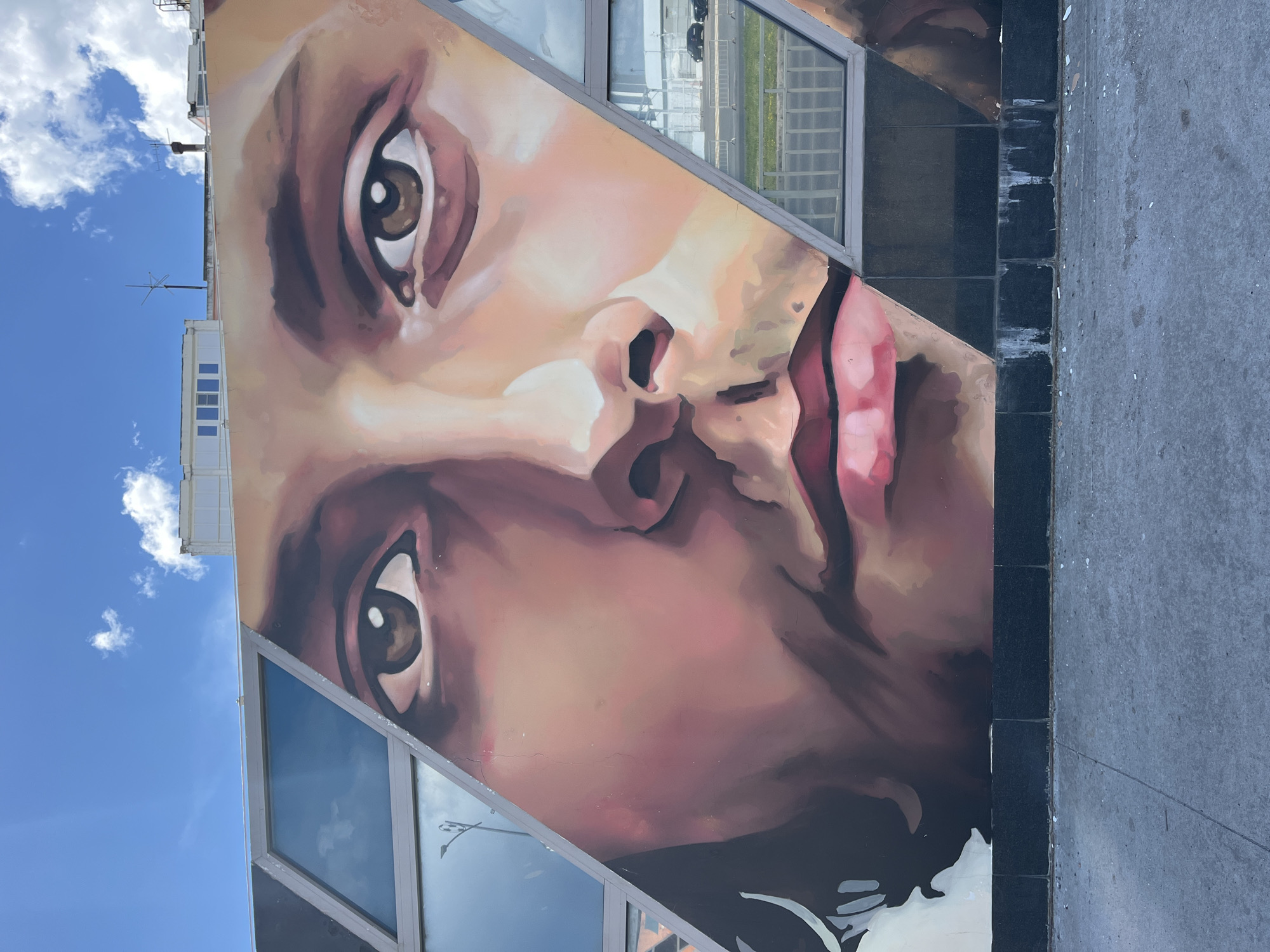 The image shows a close-up mural of a person's face painted on a building. The mural is divided into sections by diagonal lines, with windows incorporated into the design. The person's eyes, nose, and lips are prominently featured. The mural is painted in a realistic style with warm tones. In the background, there are blue skies with scattered white clouds and some buildings. Below the mural is a grey concrete sidewalk. The image is taken from a low angle, looking up at the mural. The building appears to be modern, with large windows. The mural is located in the city of Nizhny Novgorod, Russia, on a building along a street with a low fence and some cars visible through the windows.