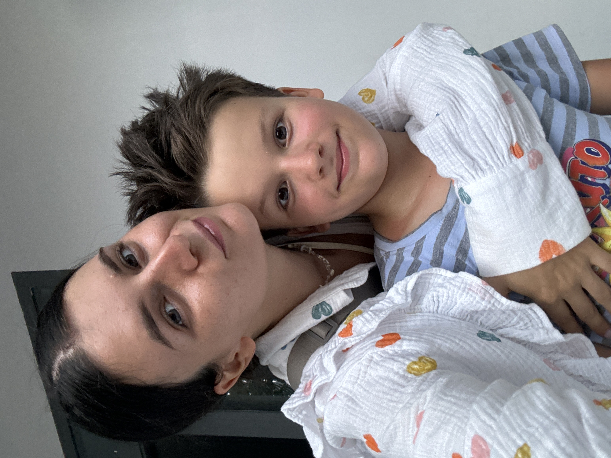 A woman and a young boy are lying down next to each other, facing the camera. The boy has short brown hair and is wearing a white shirt with hearts. The woman has dark hair pulled back and is wearing a similar white shirt. They appear to be in an indoor setting with a window or light source nearby. The specific location of Mũi Né, Vietnam cannot be determined from the image.