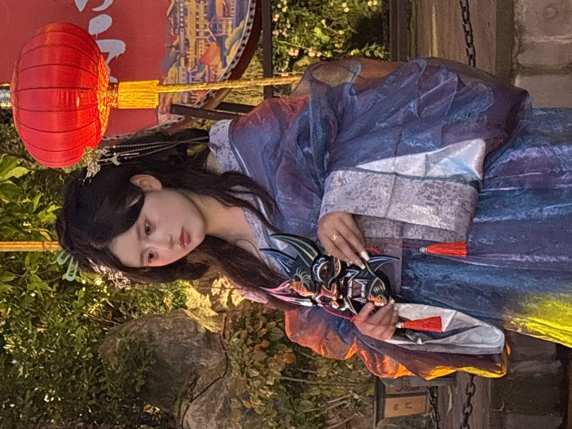 A young Asian woman is standing outside with a red lantern to her left, holding a mask. She wears a blue and grey traditional Chinese dress. There are green plants and a rock behind her. The location appears to be in Yuzhong District, China.