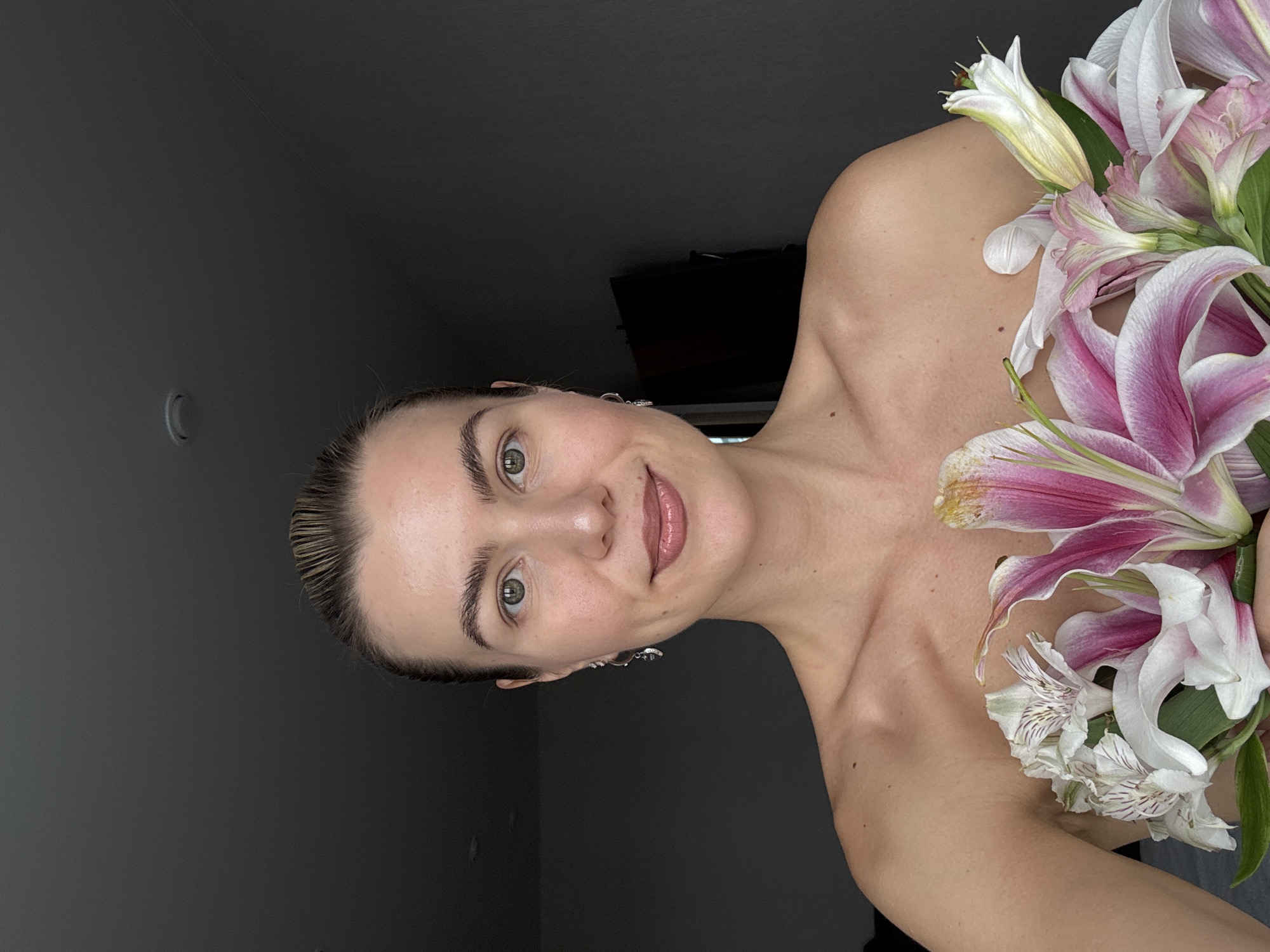 A portrait of a fair-skinned woman with light brown hair slicked back, holding a bouquet of white and pink lilies, is presented in an indoor setting. The woman's face is in focus, revealing light eye makeup and a glossy lip. She wears dangling earrings. The background is divided into shades of gray, suggesting a room with varying light levels. The flowers are positioned near her shoulder and upper chest, and their vibrant colors contrast with the more subdued tones of her skin and the background.