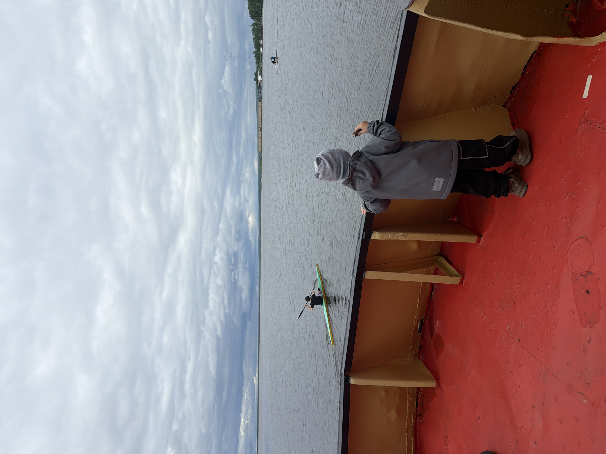 A child in a gray jacket and hood stands on a red surface and leans against a dark ledge, looking out over a body of water. A kayaker is visible in the water, with trees and buildings lining the far shore under a cloudy sky. The photo appears to be taken from a boat or dock. This image was taken in the city of Chaikovsky, Russia.