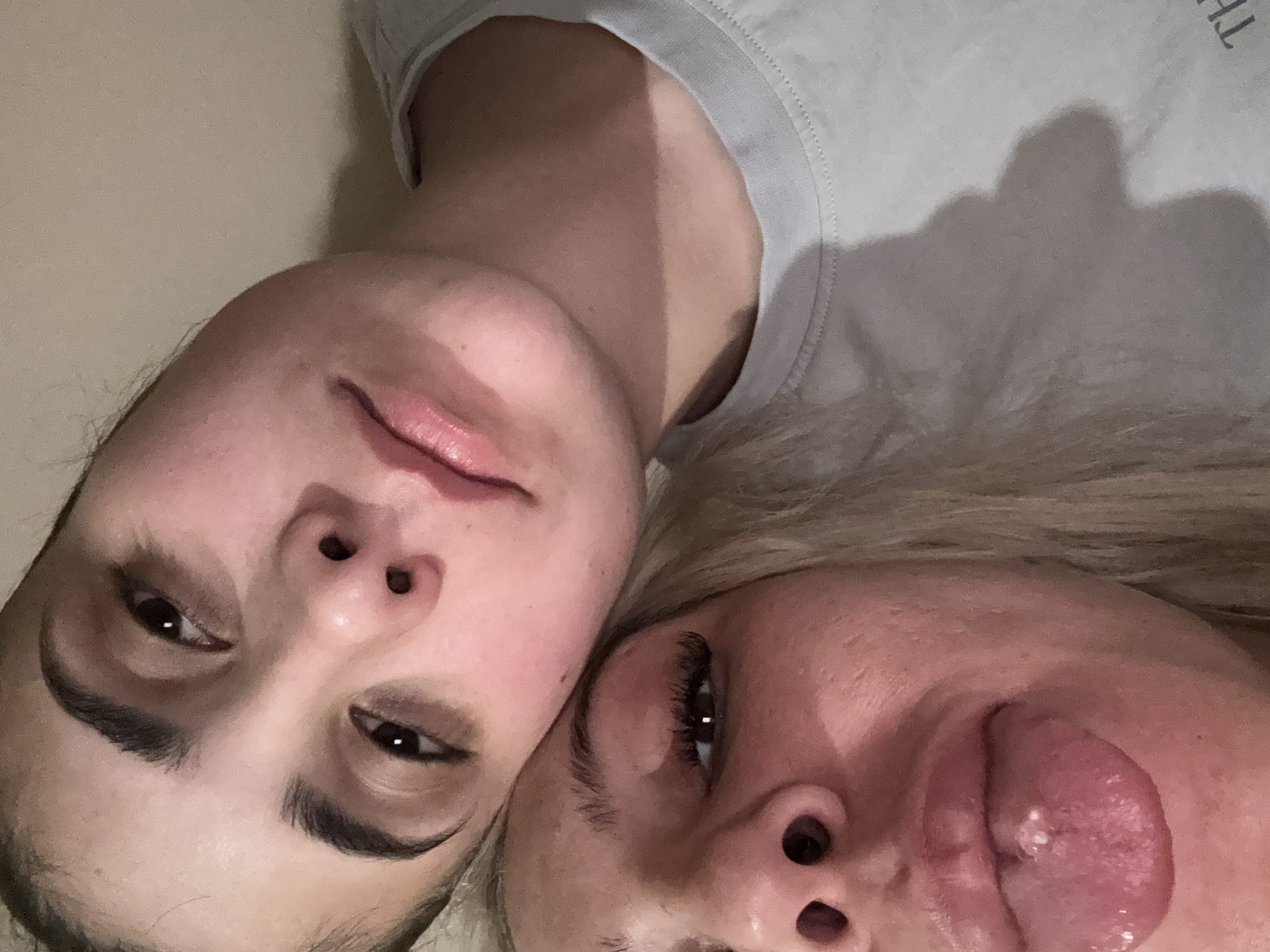 Two individuals are visible in the image. The person on the left has dark hair and is wearing a gray shirt. The person on the right has blonde hair and is sticking out their tongue. The location is Unknown, Unknown.