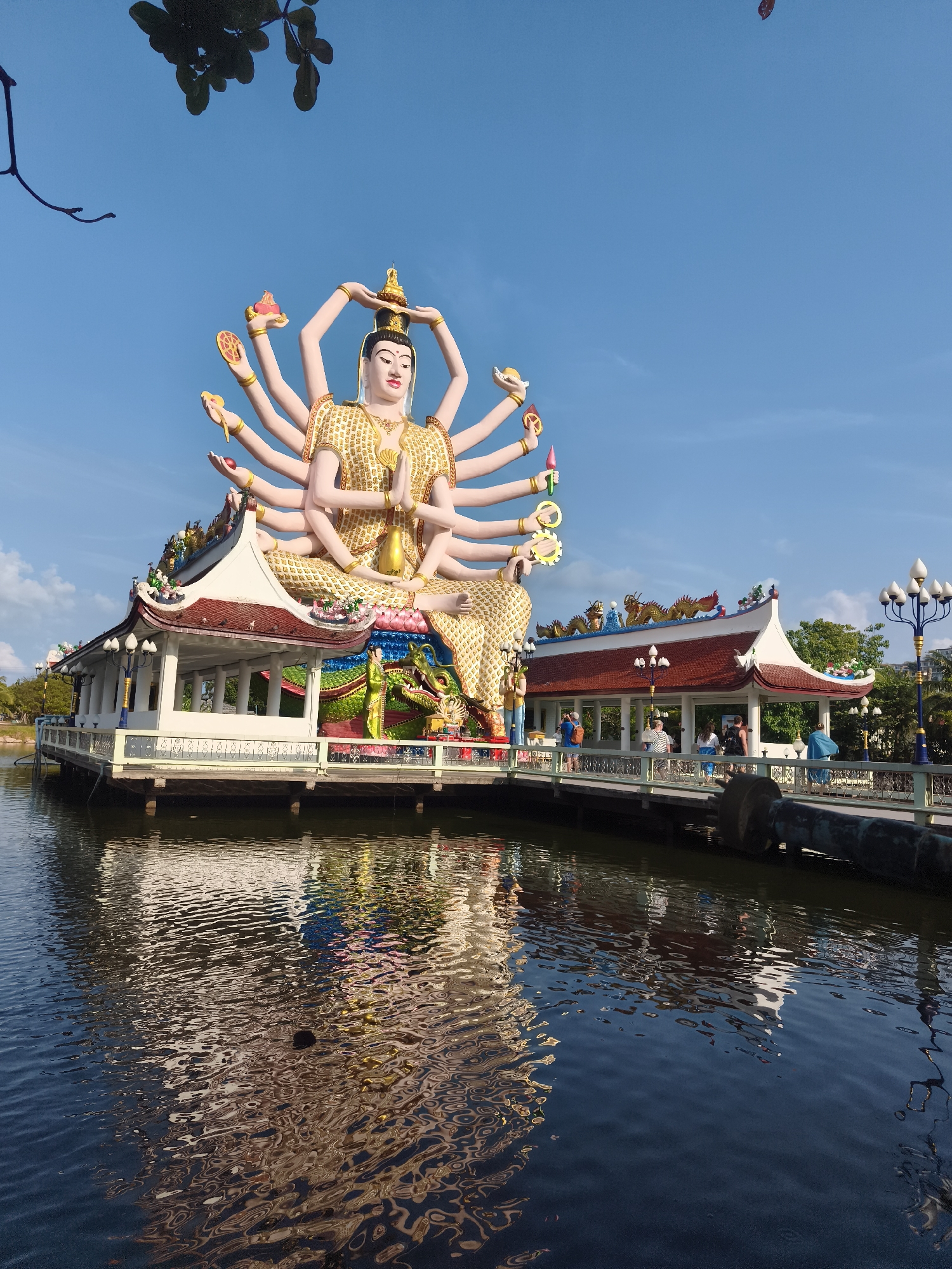 The image depicts the statue of Guanyin at Wat Plai Laem, Baan Plai Laem, Thailand. The multi-armed, seated statue is gold and pink, and is positioned on a platform above a body of water, which reflects the statue and its supporting structure. The platform is accessible via a bridge and appears to be part of a temple complex. A small group of people can be seen on the bridge and around the temple buildings. The sky is a clear blue.