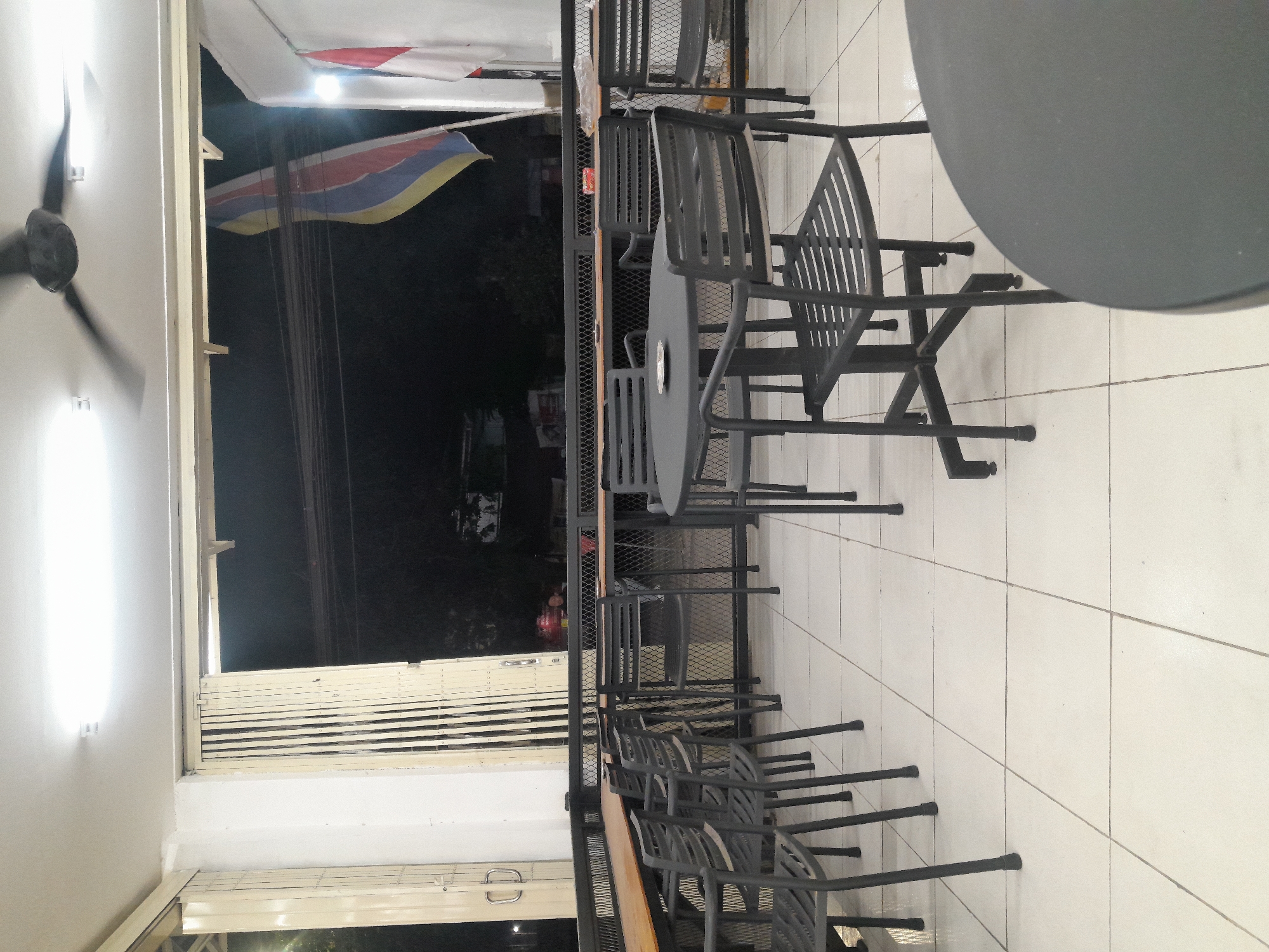The image depicts an outdoor seating area, possibly a cafe or restaurant, at night in Desa Ponokawan, Indonesia. The area is furnished with several black metal tables and chairs. The floor is tiled in a light color. In the background, a closed accordion-style gate and a partially visible flag on a flagpole are present. A ceiling fan is visible overhead, and artificial lighting illuminates the scene. The overall atmosphere suggests a quiet evening setting, with no people actively engaged in the seating area.