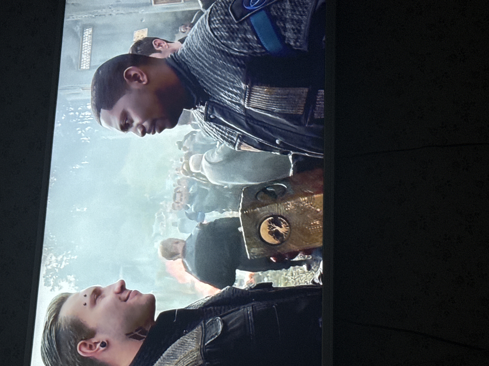 Two men stand close together against a backdrop of a crowd of people in a smoky environment. The man on the left has fair skin, light-colored hair, eyebrow piercings, and a tattoo on his neck. He is wearing a dark jacket. The man on the right has dark skin, a close haircut, and wears a uniform with a blue stripe and a patch that resembles an American flag. Between them, a person is holding a golden, boxy object with a circular design on its side. The location is not identifiable as the city of Mytishchi, Russia based on the provided image.