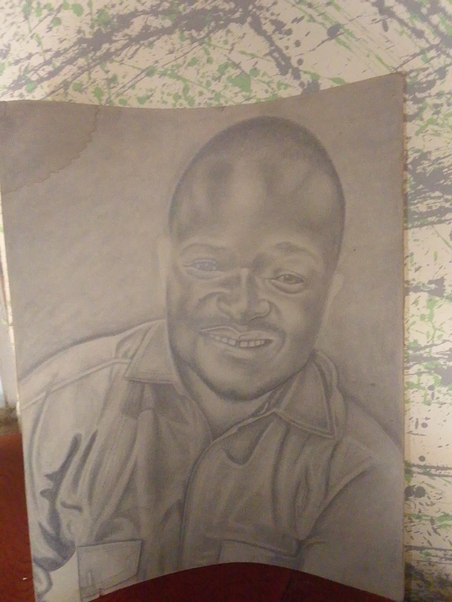 This is a graphite pencil drawing on paper, depicting a portrait of a man. The man is facing forward, with a slight smile showing his teeth. He has a prominent forehead and dark eyes. He is wearing a collared shirt, with visible folds and shadows suggesting the fabric's texture. The drawing is rendered in a realistic style, with careful attention to shading and form. The background of the drawing is a textured, abstract pattern of green and gray splatters on a light-colored surface, likely wallpaper or a similar material. This background is a stark contrast to the detailed portrait. There is a water stain visible on the upper left corner of the paper. The bottom of the drawing is partially obscured by a dark red surface, possibly furniture. The lighting in the original image appears to be natural, casting soft shadows. There is no visible text within the drawing or on the paper. The overall mood of the portrait is calm and approachable. The location context of Jalingo, Nigeria, is noted but not visually indicated within the artwork itself.