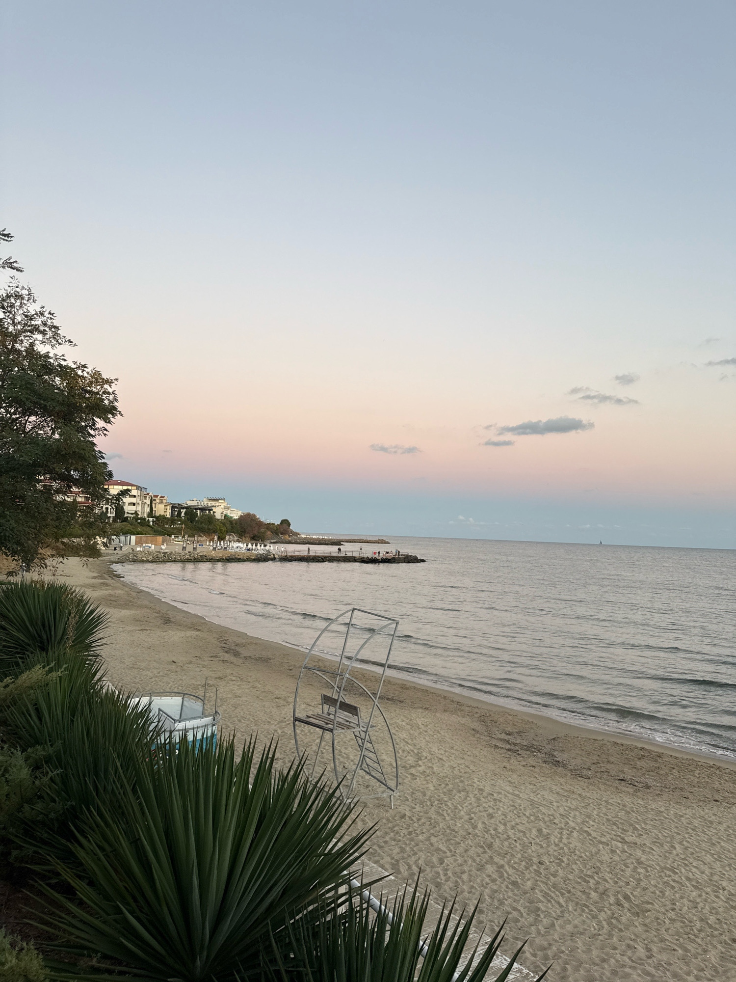 The image depicts a tranquil beach scene at dusk in Sveti Vlas, Bulgaria. The sky transitions from a soft blue at the top to hues of pink and pale orange near the horizon, indicating the setting sun. A calm sea stretches out to meet the sky, with gentle waves lapping at the sandy shore.
In the foreground, lush green spiky plants, possibly Yucca, are visible on the left, framing the beach. A metal lifeguard stand with a wooden bench is positioned on the sand. Further down the beach, a small white and blue structure, possibly a changing room or a small kiosk, is present.
The middle ground reveals a stretch of sandy beach lined with lounge chairs and umbrellas, suggesting a popular tourist destination. Beyond the beach, a row of white buildings, likely hotels or apartments, are built along a gentle slope, backed by trees. A pier extends into the sea from the right side of the buildings, with a few figures visible on it.
In the far distance, a faint outline of a sailboat can be seen on the horizon, adding a touch of serenity to the expansive sea. The overall atmosphere is peaceful and serene, characteristic of a quiet evening by the Black Sea coast.