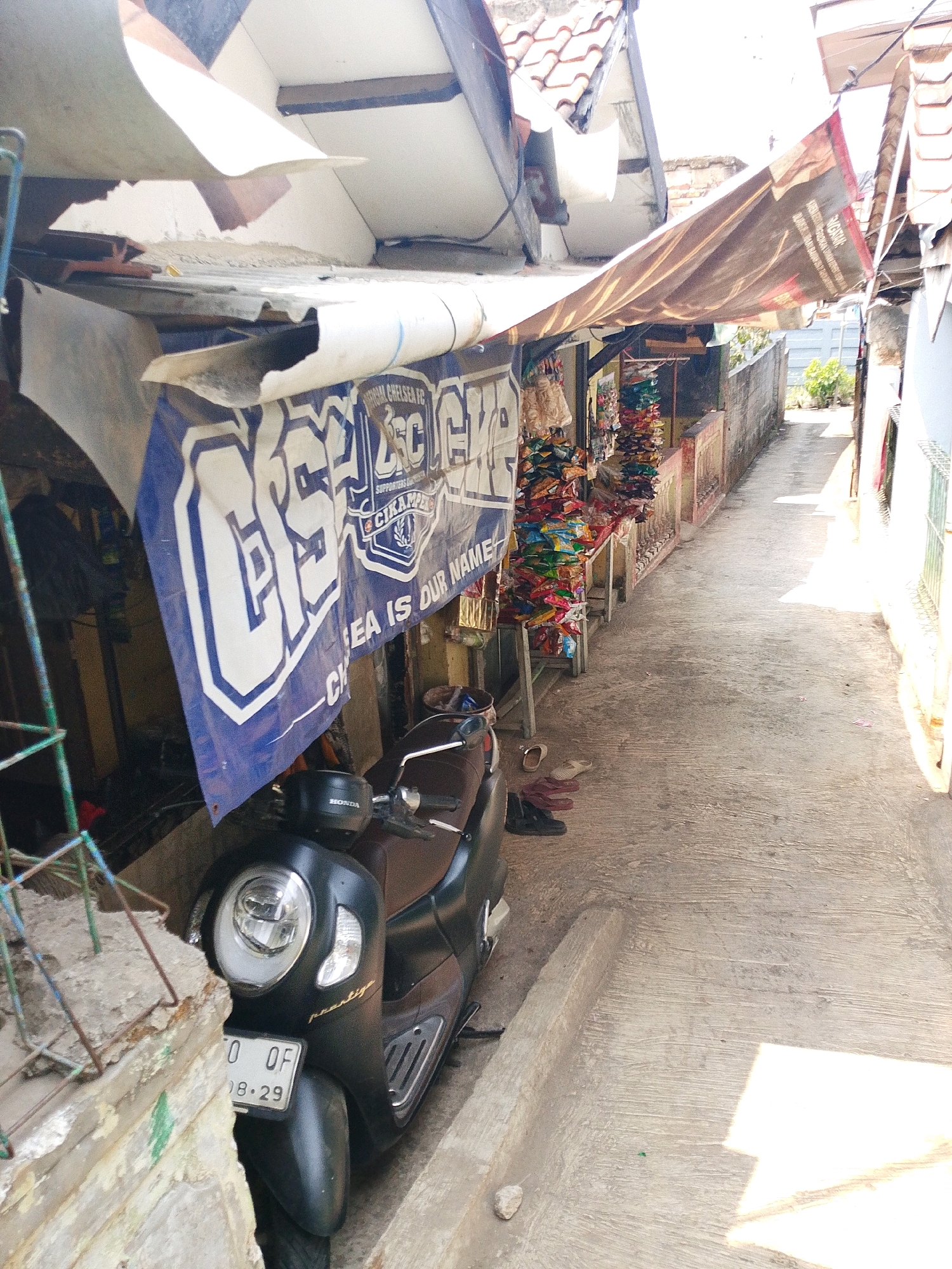 The image depicts a narrow street scene in Karawang, Indonesia. In the foreground, a black Honda scooter is parked. The scooter's license plate reads "B 0 OF 08-29". To the left of the scooter, a portion of a building's rough concrete facade is visible, with some green markings.
A large blue and white banner hangs from the overhang of a structure on the left, partially obscuring the view into the building. The banner features text in white, including what appears to be "CHELSEA FC" and "SUPPORTERS CIKAMPEK".
Along the right side of the street, a row of small stalls or shops is lined up. These stalls display a variety of colorful merchandise, predominantly bags of snacks. The stalls are covered by a brown tarp. The street itself is paved with concrete, with visible expansion joints and some scattered debris, including a few pairs of sandals and shoes. The environment suggests a commercial or residential alleyway, with buildings and awnings lining both sides, and a bright, sunny day. The overall impression is of a vibrant, everyday street scene in a Indonesian city.