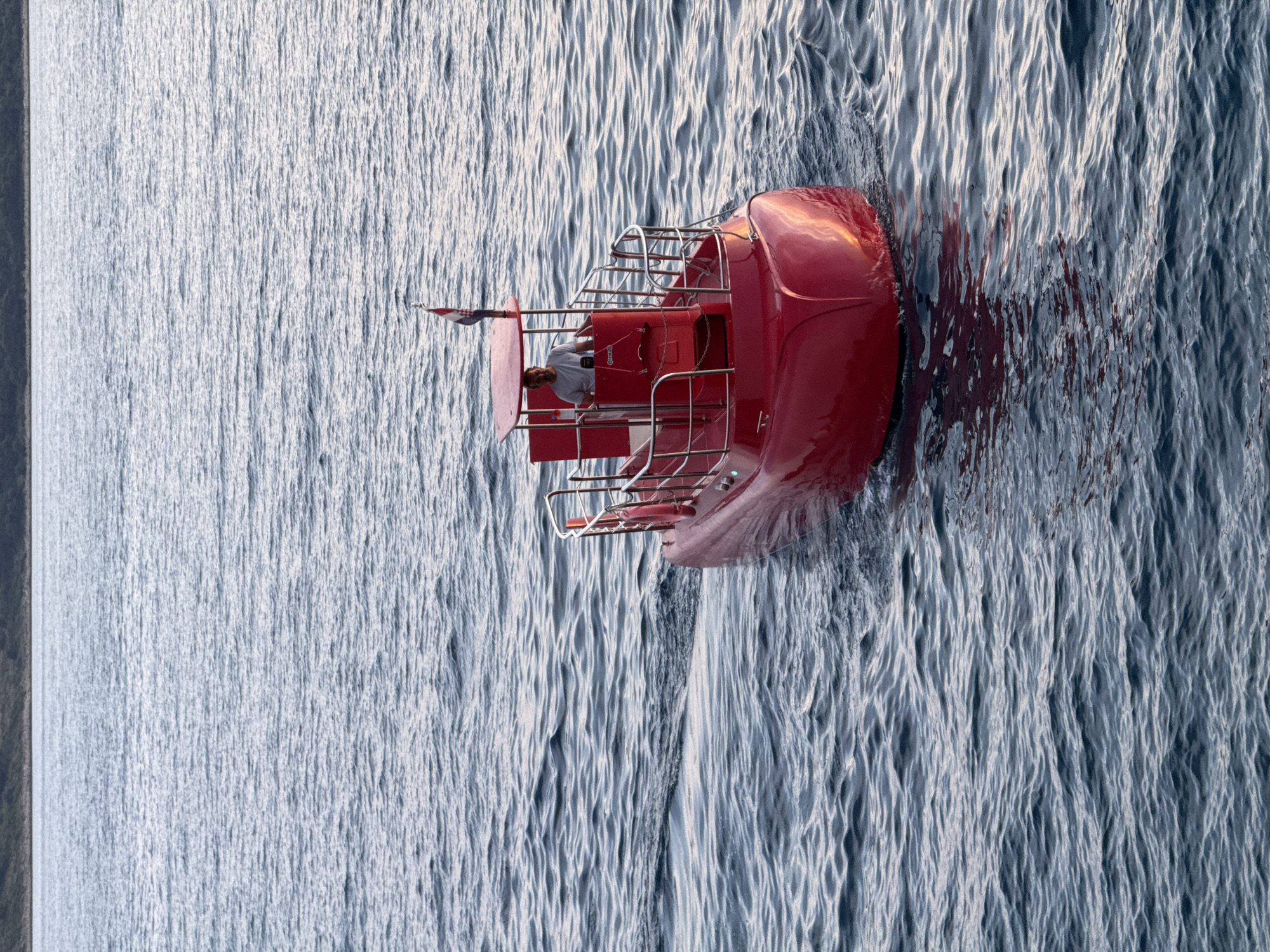 The image shows a bright red, small passenger submarine, or semi-submarine, with a clear canopy, navigating through calm, blue-grey water. A man, wearing a light-colored t-shirt, is visible at the helm of the submarine, steering the vessel. A flag, appearing to be the flag of Croatia, is planted on the rear of the submarine's roof. The submarine is creating a wake as it moves. In the background, a dark, low-lying coastline with vegetation is visible on the horizon. The lighting suggests it is either early morning or late afternoon, with a soft, warm light reflecting on the water and the submarine. This scene is characteristic of the waters surrounding Primošten, Croatia, a coastal town known for its clear waters and tourist activities.