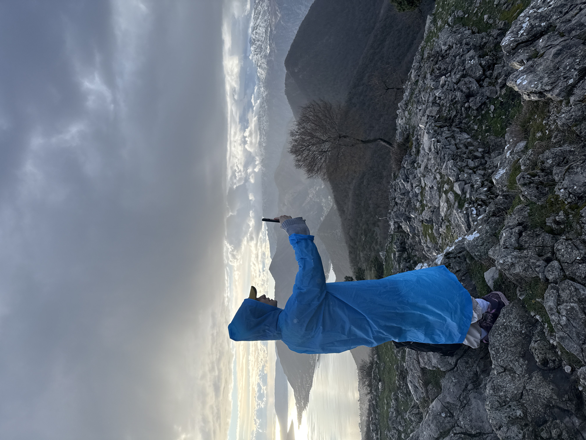 A sole individual stands on a rocky, uneven mountain ridge at an elevated position in Bogdašići, Montenegro, overlooking a vast panoramic landscape. The person, viewed from a slightly elevated rear-left angle, wears a bright blue hooded poncho, a yellow baseball cap, tan pants, white socks, and purple hiking shoes. A black backpack is partially visible behind them. They are actively holding a black smartphone vertically with both hands, extended forward, seemingly capturing the expansive view. To the left, a large, calm body of water, resembling a bay or fjord, reflects bright sunlight breaking through an overcast sky. Coastal settlements are visible along its shores. The midground and background consist of multiple layers of dark, rugged mountains. A solitary, bare deciduous tree with a contorted trunk stands on a ridge to the right of the individual. Distant mountain peaks in the far background are snow-capped under a heavy, dark grey cloud cover. The foreground is composed of large, grey rocks with sparse green vegetation and white lichen patches.