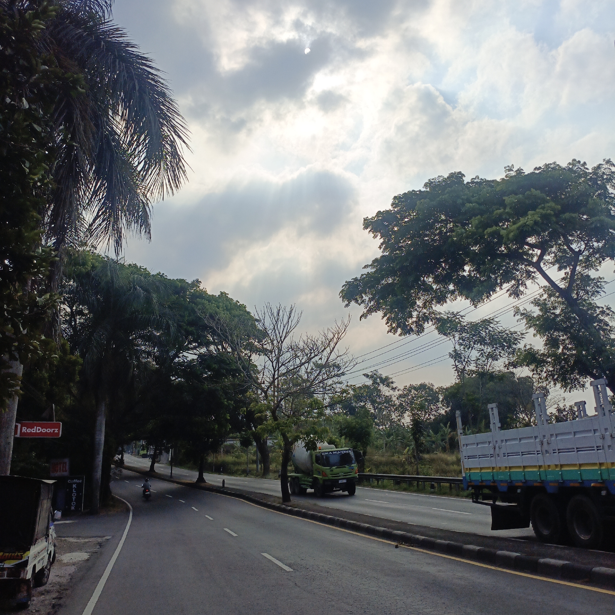The image depicts a street scene in Salatiga, Indonesia, on a cloudy day with sunlight breaking through. On the left side of the road, a RedDoorZ hotel sign is visible above a building with a sign for "Coffee KLOTOK". A truck is parked on the side of the road near the hotel, and a motorcycle with a rider is moving away from the camera on the road. The road itself is paved with dashed white lines. In the middle ground, a green cement mixer truck drives on the road. On the right side, a flatbed truck with a blue, green, and yellow stripe design travels on the road. Tall trees line the sides of the road, with dense foliage visible. Power lines are strung across the background. The sky is filled with dramatic, backlit clouds.