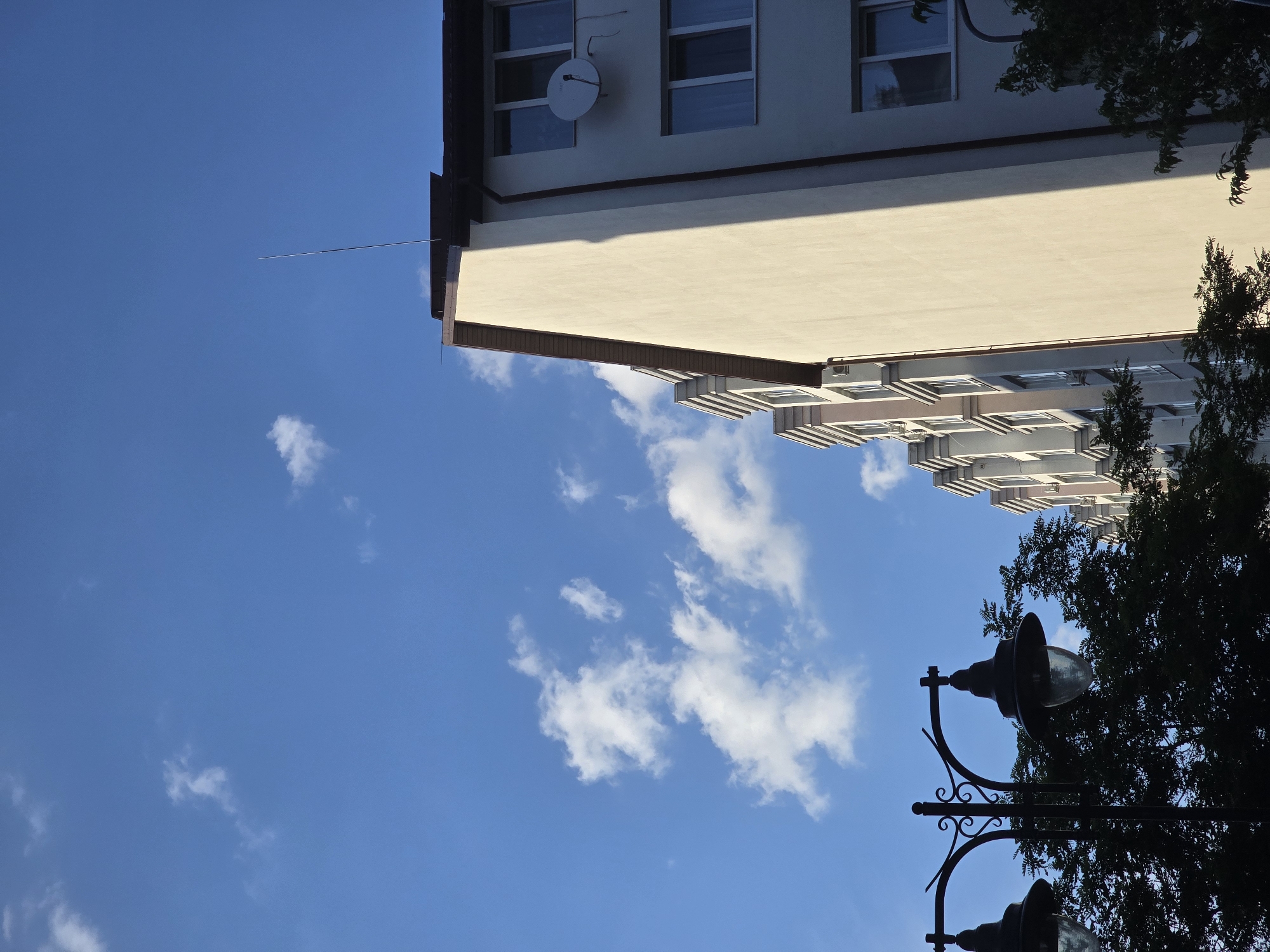 The image depicts a bright, sunny day with a clear blue sky and scattered white clouds. The perspective is skewed, making it appear as if the viewer is looking upwards. In the foreground, silhouetted against the sky, are two ornate lampposts with decorative scrollwork. Dense, dark green foliage from a tree partially obscures the lampposts.
Dominating the upper portion of the frame is the side of a building. The building has a light-colored facade, with a section in shadow cast by the building itself. Several windows are visible, some reflecting the sky. A satellite dish is mounted on the wall of the upper section of the building. Extending from the building's roof is a thin, metallic rod.
The scene suggests an outdoor urban or suburban setting, possibly a street or park area adjacent to residential buildings. The sunlight indicates it is daytime, likely midday or afternoon given the angle of the shadows. The weather appears pleasant and clear. No people are visible in the image. There is no visible text.