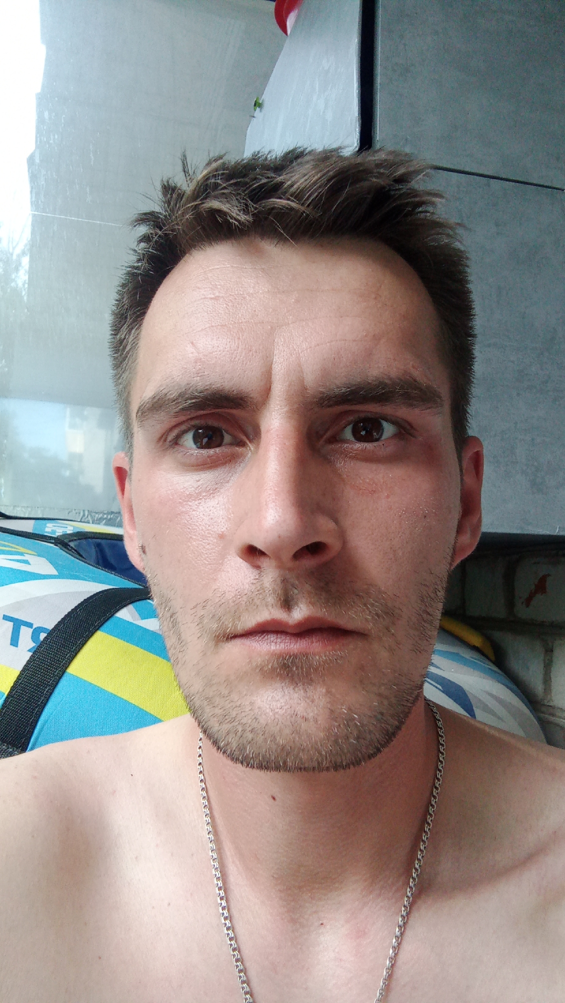 The media file is a close-up selfie of a man's face and upper chest. He has short, messy brown hair and a light stubble beard. His skin appears slightly oily, and there are some visible wrinkles on his forehead and around his eyes. He is wearing a silver chain necklace.
The man's expression is neutral, and his eyes are looking directly at the camera. The background is blurred but appears to be indoors, with a hint of blue and yellow fabric in the lower left corner and a grey, textured wall behind him. There is also a red object visible in the upper right corner of the frame. The lighting suggests it might be daytime, possibly overcast, given the lack of strong shadows.
The context provided indicates the location is Chaikovsky, Russia, but there are no overt visual cues in the image to confirm this. No text is visible in the image. The overall impression is a candid, unposed shot of the man.