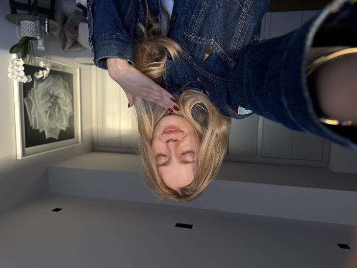 An indoor, eye-level selfie captures a blonde female, approximately 20-30 years old, with her eyes closed and a subtle smile. She wears a blue denim jacket featuring light-colored stitching and a partial patch on the left shoulder. Her left hand, adorned with dark red nail polish and a ring on the ring finger, rests gently on the right side of her neck/collarbone. A gold-toned bracelet or watch is visible on her right wrist, which is extended towards the camera. The background features light-colored walls and ceiling with recessed lighting. To the right, a large framed black and white artwork depicting a rose hangs above a small, dark round table holding a white orchid plant and several small bottles. Further left, a set of white doors or cabinet panels are visible. Strong natural light illuminates the scene from the right, creating warm highlights. The setting is a room in Mexico City, Mexico.