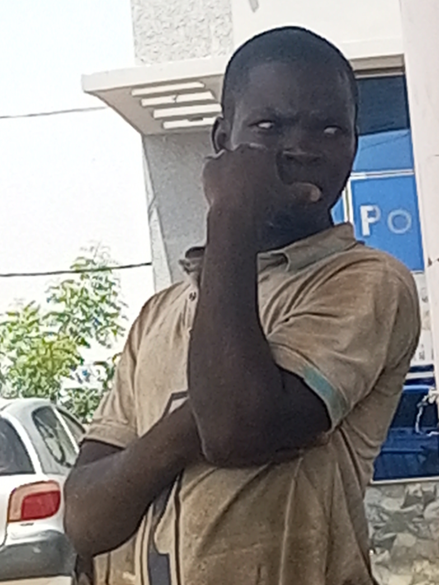 A person with dark skin and white eyes is standing in front of a building. The person is wearing a tan short-sleeved polo shirt and has their arms crossed with one hand near their face. A car is partially visible in the background, and some trees are to the left. A sign with the letters "PO" is visible in the background near the person. The location is inferred to be Gombe, Nigeria, based on prior knowledge.