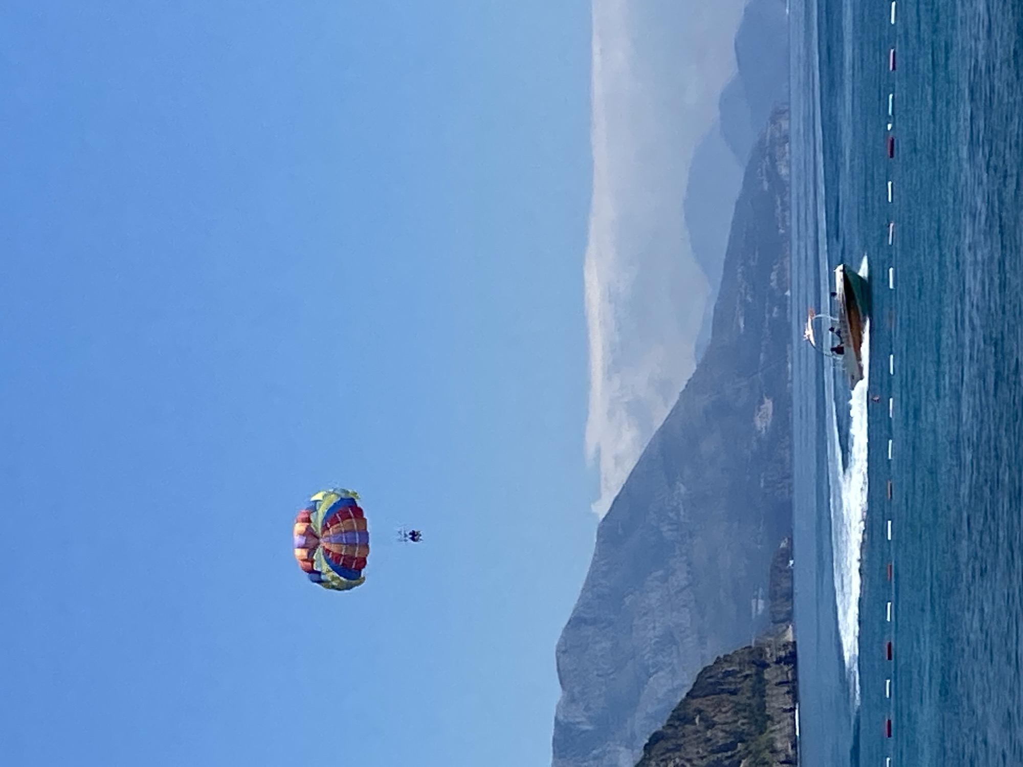 In the city of Prijevor, Montenegro, on a clear day with a vibrant blue sky, a parasailer drifts above a tranquil blue sea. The parasail, a colorful canopy of red, yellow, blue, and green, is tethered to a boat that speeds across the water, leaving a white wake. Two figures are visible, suspended beneath the parasail, enjoying the aerial view. In the background, a series of majestic, layered mountains rise from the coastline, their peaks softened by a hazy, white cloud formation hugging their upper slopes. The foreground of the scene is marked by a floating barrier of red and white buoys, delineating a boundary in the water.