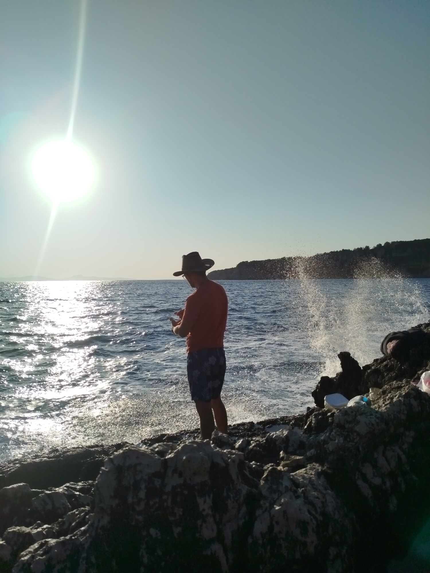 A person stands on rocky terrain by the sea in Karaburun, Turkey, facing away from the camera and towards the water. The person is wearing a hat, an orange t-shirt, and patterned shorts. They are holding a mobile device in their hands and appear to be looking at it. The sun is bright and low in the sky, casting a strong glare on the water. Waves are crashing against the rocks, creating a spray. The background shows a coastline with trees.