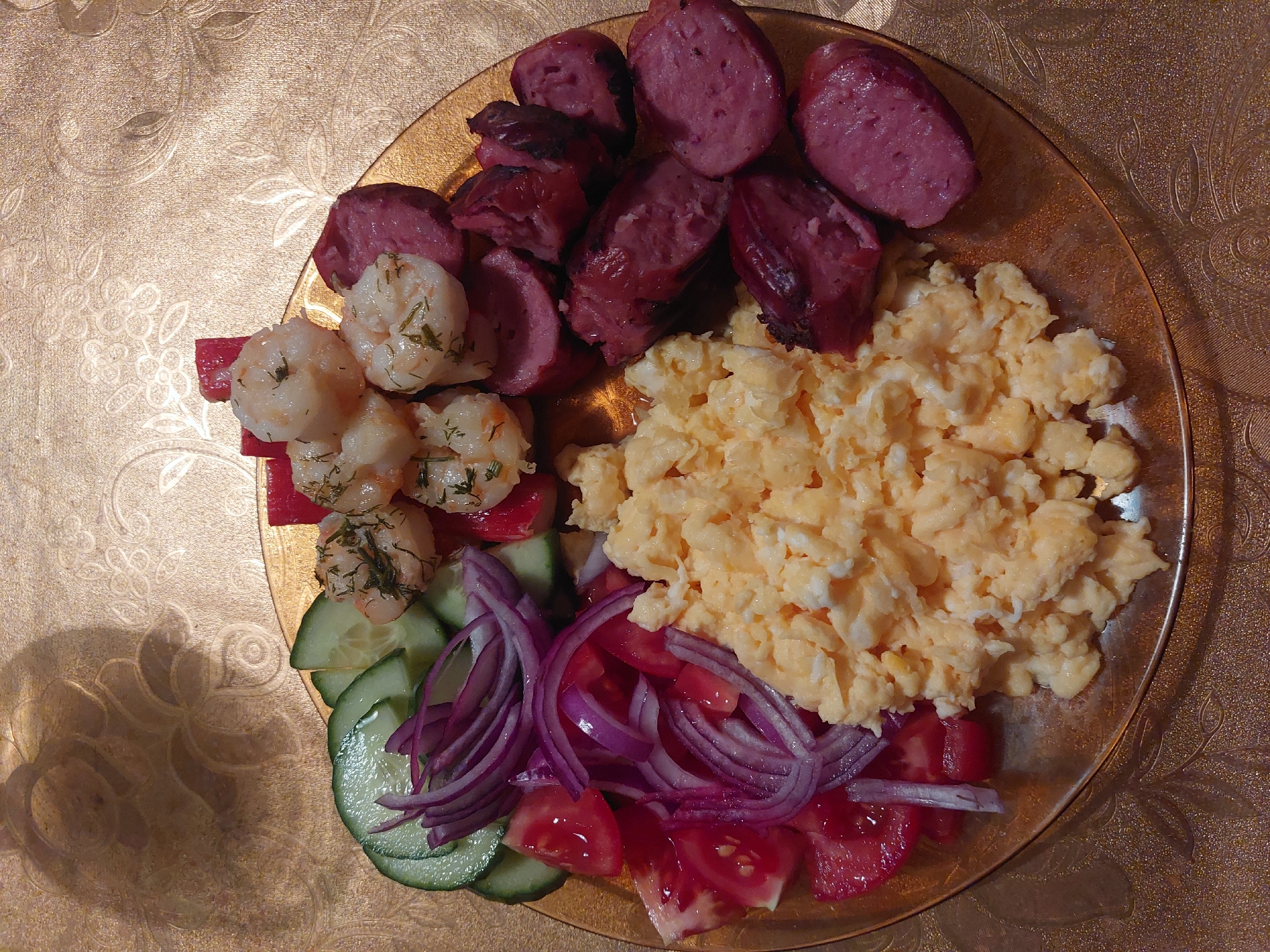 The image shows a plate of food. The plate contains sliced sausage, scrambled eggs, what appear to be shrimp mixed with dill, cucumber slices, red onion slices, and tomato wedges. The plate sits on a gold-colored surface with a floral pattern. There is no evidence to identify the location as Tsarevo, Bulgaria. The image contains no people or actions.