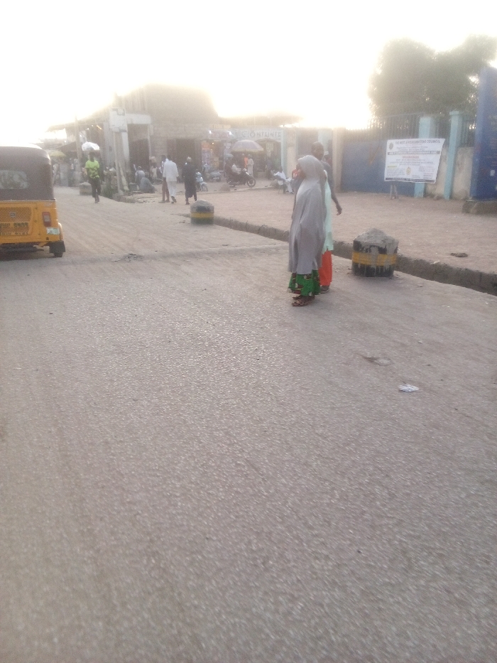 This image captures a street scene in Dakata, Nigeria, characterized by daily activity under bright, hazy sunlight, suggesting either early morning or late afternoon.
The foreground is dominated by a gray asphalt road. Two concrete-filled tire barriers, painted with yellow and black stripes, are positioned on the right side of the road, acting as dividers or bollards. A similar barrier is visible further back on the left.
In the midground, several people are present. Two women stand on the right, with their backs to the camera; one wears a light-colored head covering and garment over a green patterned skirt, and the other has a light green top with orange trousers. Further back, a small gathering of people congregates near a row of shops. One individual in a light green shirt is seen walking on the left side of the street. A yellow motorized tricycle (auto-rickshaw) is parked on the far left.
The background shows a commercial area on the left, with various shops or market stalls and more people moving about. On the right, a blue gate leads to a building, likely a public or community facility, where a large white sign is mounted. Clearly visible text on this sign reads "DANGE LOCAL GOVERNMENT COUNCIL," indicating a local administrative presence. Other text on the sign is illegible due to distance and resolution, but pertains to official information. Faintly visible text on a shop building in the left background appears to include "CONTAINER." The scene conveys a typical day in a bustling local street environment.
