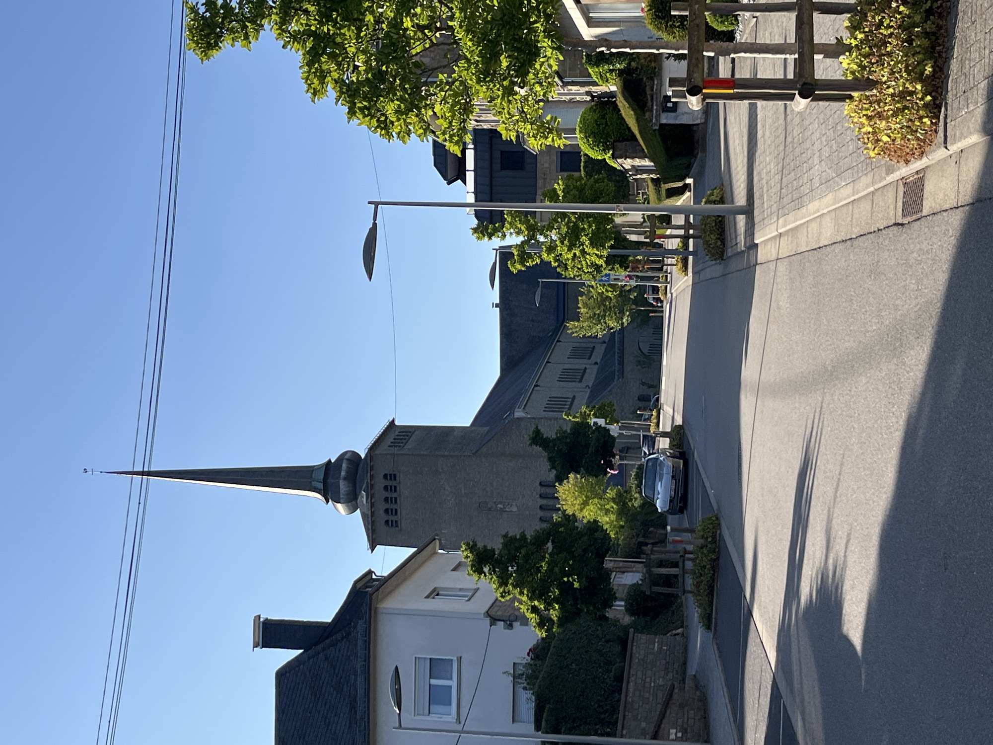 The image depicts a street scene in Saint-Vith, Belgium, under a clear blue sky. A tall church spire, characteristic of the town's architecture, dominates the background, with its dark, pointed roof and segmented tower. The street is paved, with a sidewalk lined with cobblestones and interspersed with small trees and shrubs in wooden planters. On the left side of the street, a white house with dark shutters and a dark-tiled roof is visible, partially obscured by trees. Further down the street, several other buildings, including a larger stone structure that appears to be part of the church complex, are present. A silver car is parked on the side of the road. The scene is bathed in sunlight, casting long shadows across the street. Electrical wires are strung across the top of the image. The overall impression is one of a quiet, residential street in a European town.
