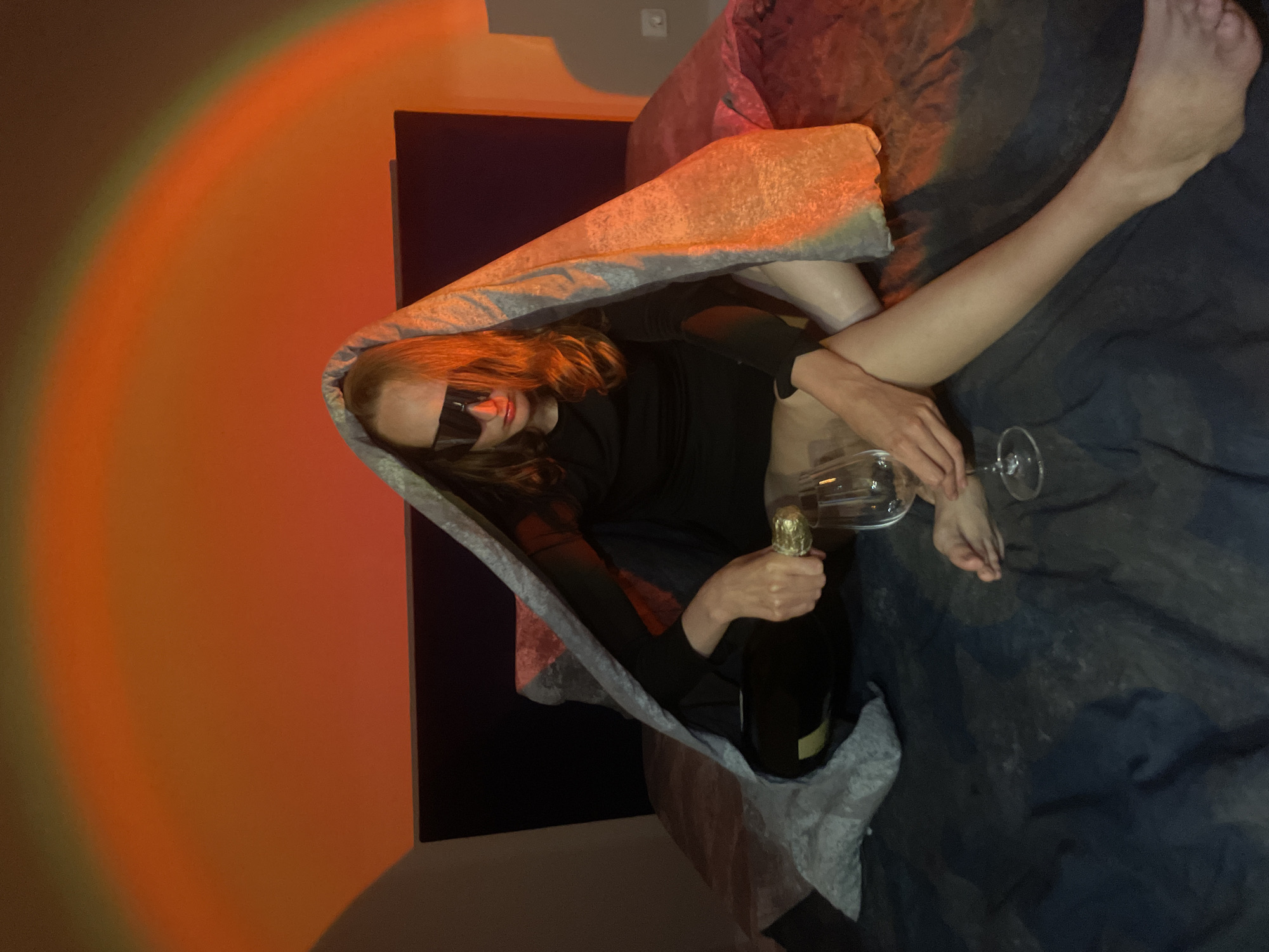 A woman is lying on a blue textured surface, possibly a bed, covered partially by a textured blanket. She wears a black top and dark sunglasses, holding a bottle of sparkling wine in one hand and a wine glass in the other. An orange light casts a glow on the adjacent wall. The location, Подшивалово, Russia, cannot be independently verified from this image.