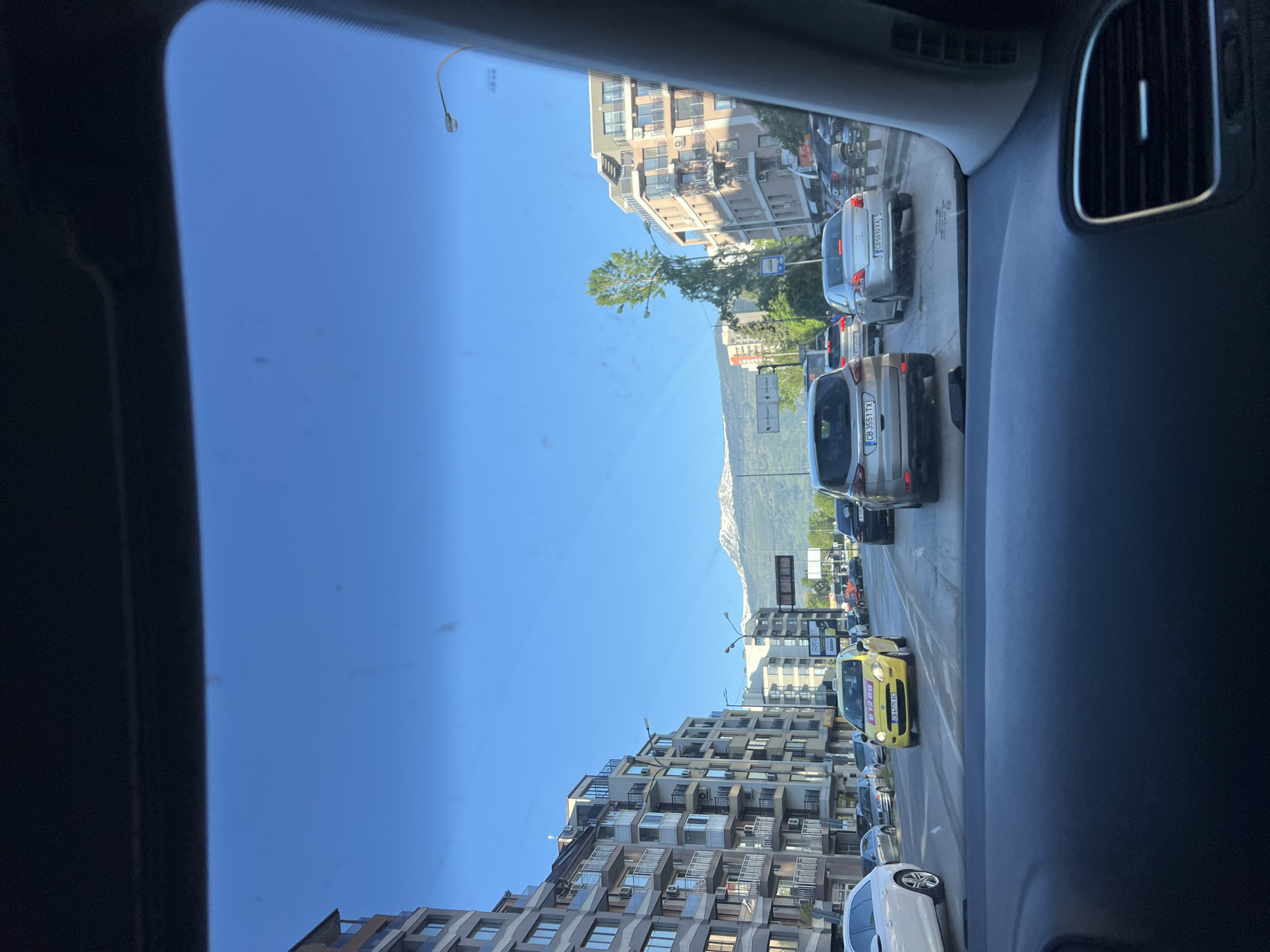 The image is taken from inside a car in traffic in Sofia, Bulgaria. The car is facing towards the Vitosha mountain, which is visible in the distance. Buildings line both sides of the street, a bright blue sky is above. Several cars are visible in front of the car, including a yellow taxi and a white car. The registration plates on some cars are visible, bearing Bulgarian plates.