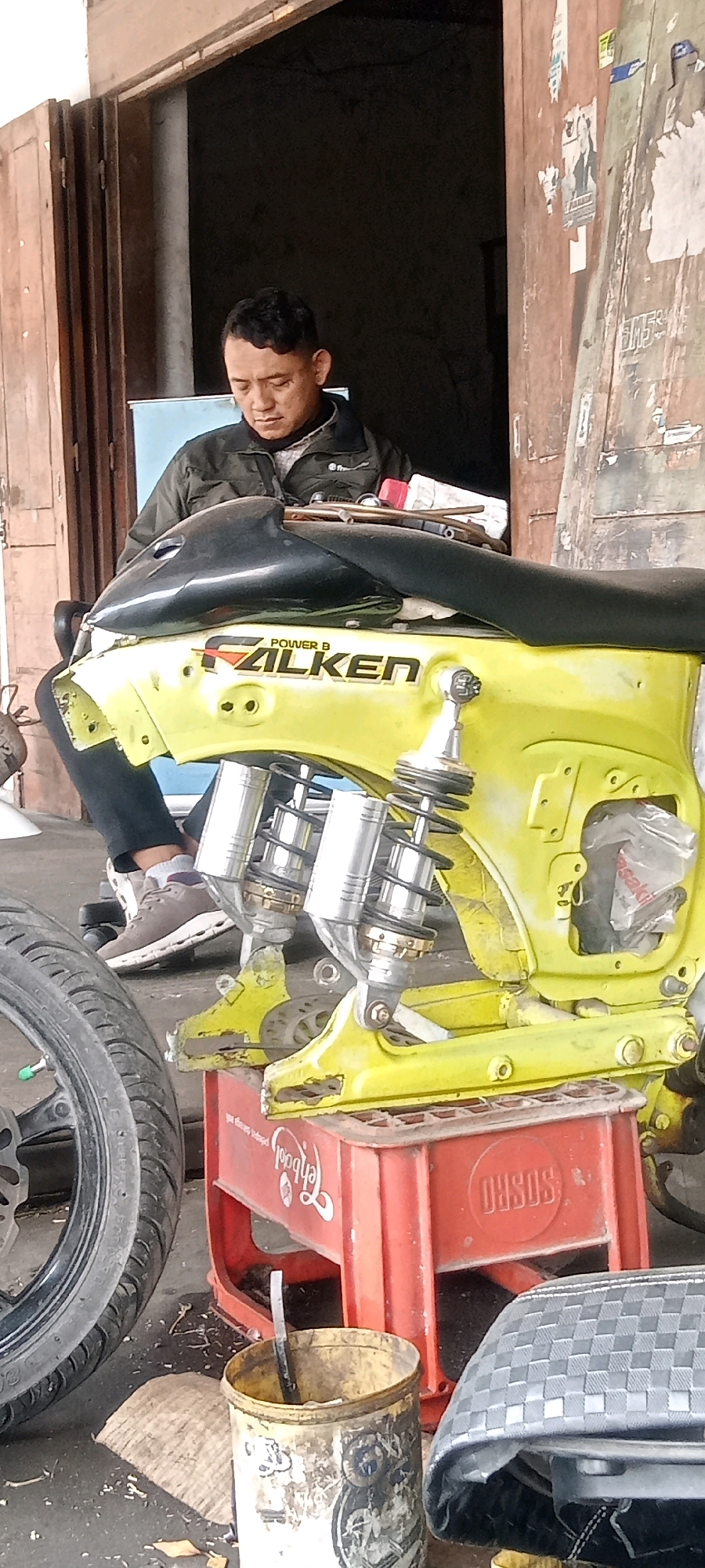 A man, appearing to be of Indonesian descent with short black hair and wearing a dark jacket over a light-colored shirt, sits in front of a bright yellow motorcycle frame with black detailing. He is looking down, possibly at the motorcycle or something in his lap. The motorcycle frame is the primary subject, featuring prominent chrome shock absorbers with black springs, and a black fuel tank. The "FALKEN POWER B" logo is visible on the yellow frame. The motorcycle is supported by a red plastic crate with white lettering, one side of which is partially obscured by the motorcycle. In the foreground, a worn tire is visible on the left, and a dirty white and yellow bucket with a tool inside sits on the right. The background consists of a wooden structure with a large open doorway, revealing a dark interior. The scene appears to be an outdoor workshop or a roadside stall, consistent with a location in Keteguhan, Indonesia. The overall environment suggests a repair or customization setting for motorcycles.