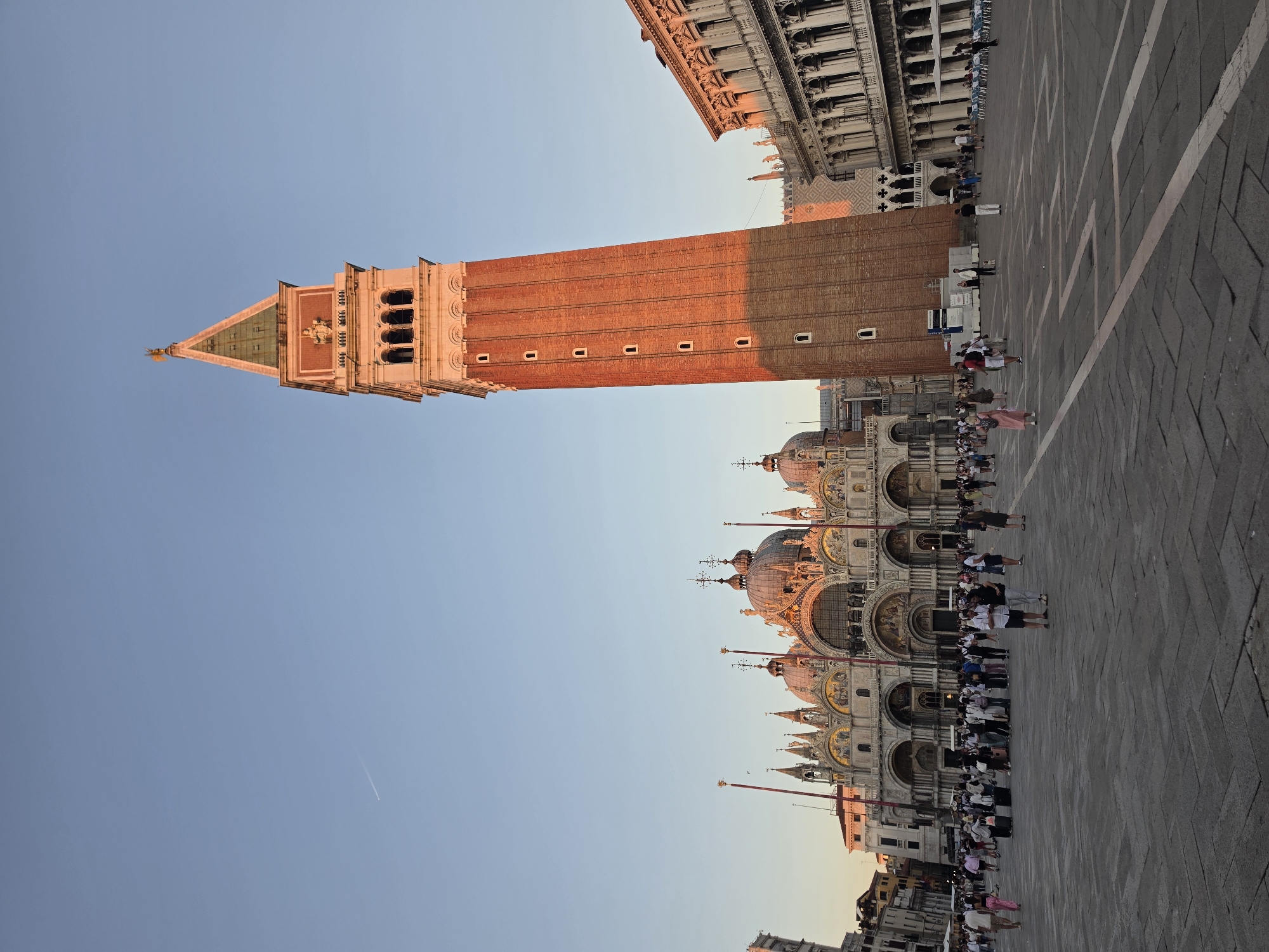 The image depicts a wide-angle view of St. Mark's Square (Piazza San Marco) in Venice, Italy, during what appears to be the late afternoon or early evening, given the warm lighting. In the background, the iconic St. Mark's Basilica, with its intricate facade and domes, is visible. To the right of the basilica stands the Campanile di San Marco, a tall brick bell tower, with its upper section bathed in sunlight. Adjacent to the Campanile is the Procuratie Nuove, a Renaissance building with arcaded ground floors.
The square itself is paved with intricate stone patterns and is populated by a moderate number of tourists. People are seen walking, standing in groups, and observing the architecture. Some individuals are carrying luggage, suggesting they might be arriving or departing. The sky is clear and a pale blue, with a faint contrail from an airplane visible in the upper left quadrant. The overall scene conveys a sense of a popular tourist destination with its historical landmarks and a bustling, yet not overcrowded, atmosphere.