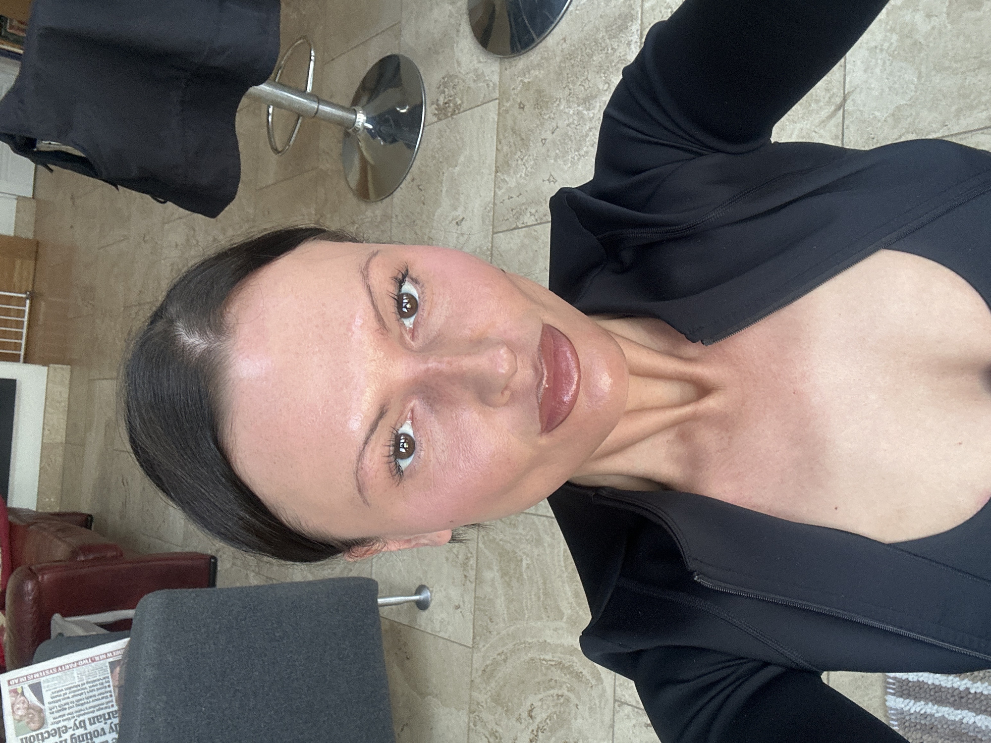 A female subject is centered, framed from the upper chest, gazing directly forward. She has dark hair pulled back, a neutral facial expression, pronounced lips with glossy reddish-brown pigment, and mascaraed eyelashes. Her facial skin exhibits a flushed appearance. She is clothed in a black, long-sleeved, zippered garment, partially unzipped. The background reveals an indoor tiled floor of light, patterned stone. To the subject's left, a dark grey upholstered chair arm is present, upon which a newspaper is visible, displaying English text including "arian by-election" and "PARTY SYSTEM IS READ." A reddish-brown leather armchair is partially discernible behind the grey chair. To the subject's right, a black textile item is draped over a silver bar stool featuring a circular base; a second similar stool base is partially visible further right. A segment of a light-colored interior wall or gate is observable in the upper right background. The scene is located within a domestic or commercial interior in the city of Wealden, United Kingdom.