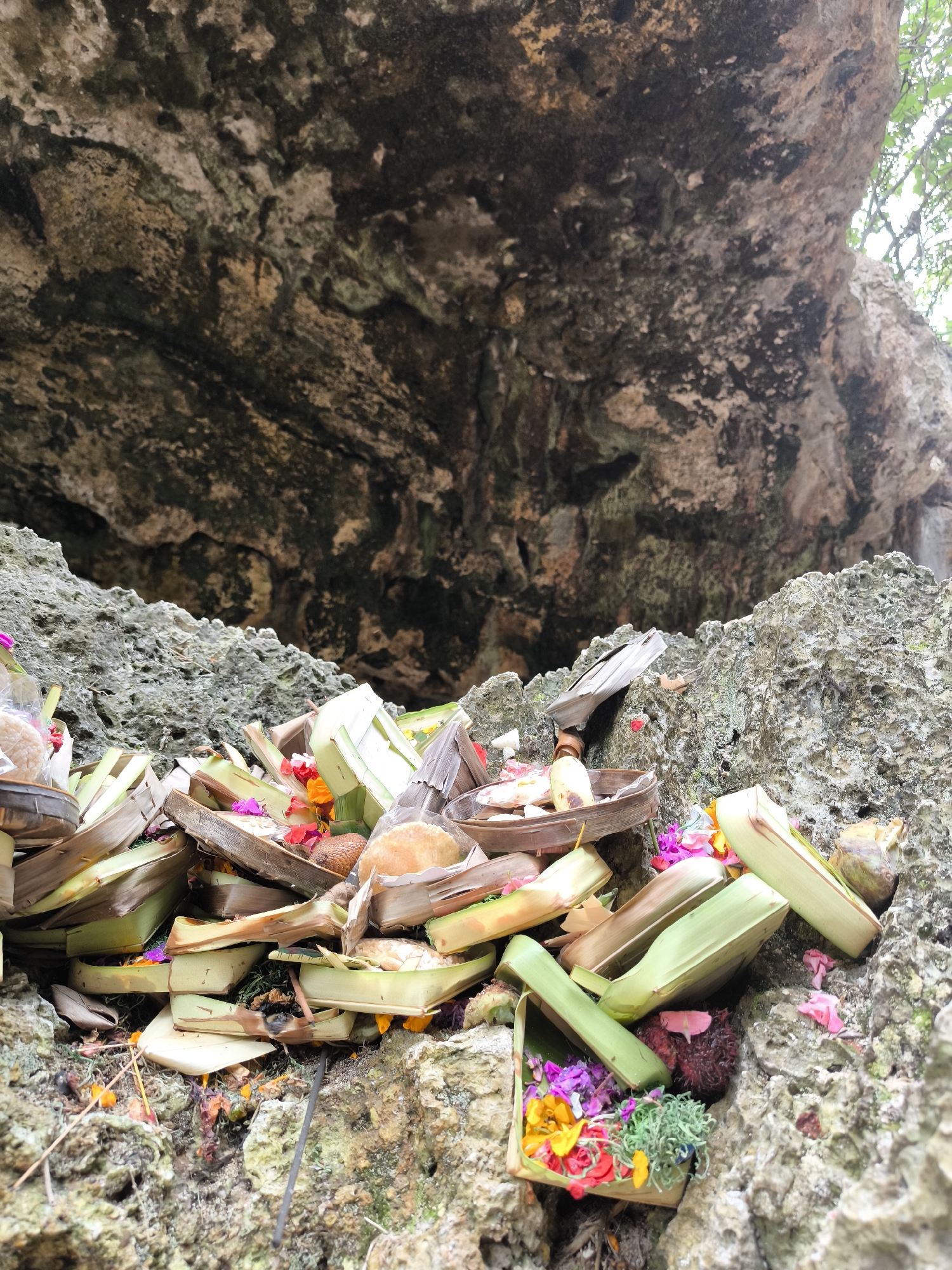 A close-up image shows Balinese offerings placed on a rocky surface beneath a rocky overhang, likely a cave formation, at Uluwatu, Indonesia. The offerings are presented in small, woven containers made from palm leaves and other natural materials. They are filled with colorful flowers, fruits, and other items. The offerings are densely packed together. The rock surface has a textured appearance. The cave formation behind the offerings is dark and has various cracks and crevices. Some green leaves are visible at the top right of the image.