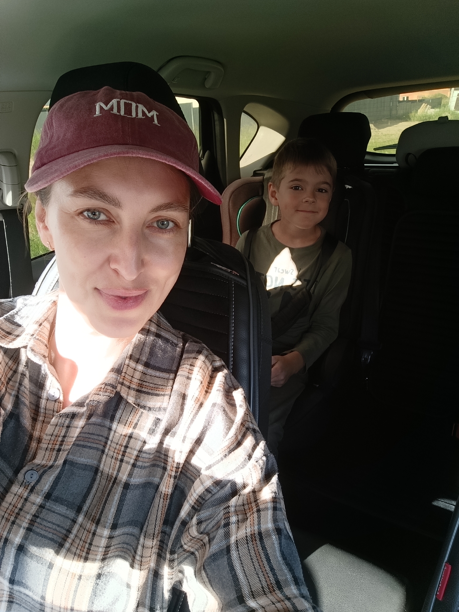 In the foreground of the image, a woman wearing a plaid shirt and a "MOM" baseball cap is taking a selfie. Her hair is pulled back, and she is looking at the camera with a slight smile. Sunlight streams in from the side, illuminating her face. In the background, a young boy is seated in a car seat. He is wearing a green long-sleeved shirt and looking towards the camera. The interior of the car is visible, with car seats and windows showing an outdoor environment. The scene appears to be in a residential area.