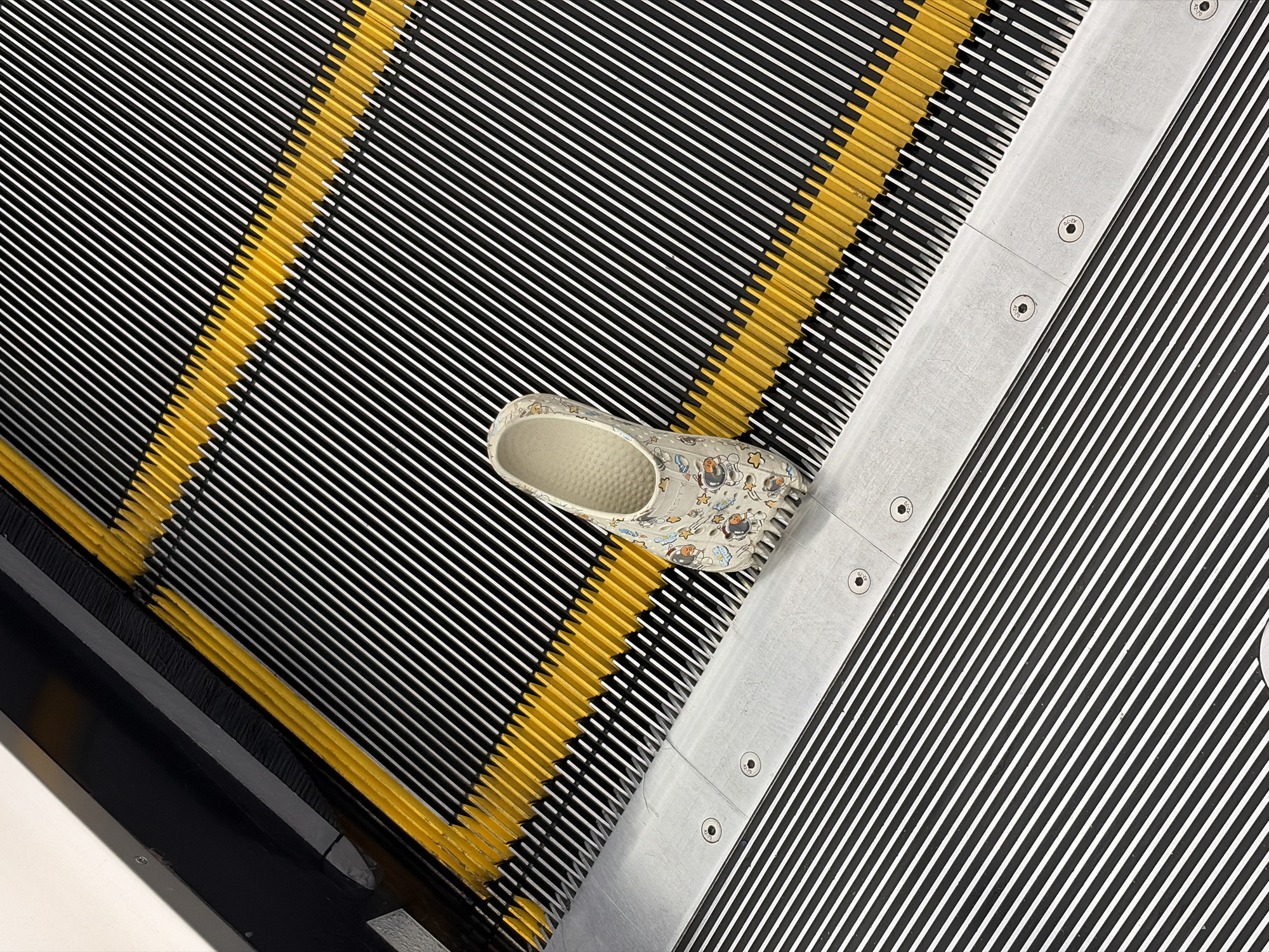 A single light-colored sandal, patterned with cartoon astronauts and stars, lies on the ribbed surface of an escalator step in the city of Thủ Đức, Vietnam. The escalator steps are predominantly black with narrow white horizontal grooves. A distinct diagonal yellow line, composed of interlocking comb teeth, delineates the boundary between the moving steps and a fixed silver-grey metal comb plate. This comb plate features four visible circular fasteners and parallel grooves that align with the escalator steps. The sandal is positioned towards the bottom right of the image, resting on a black step with its heel near the comb plate. A black escalator skirt panel with a grey top edge is visible along the upper left side. No people are present.