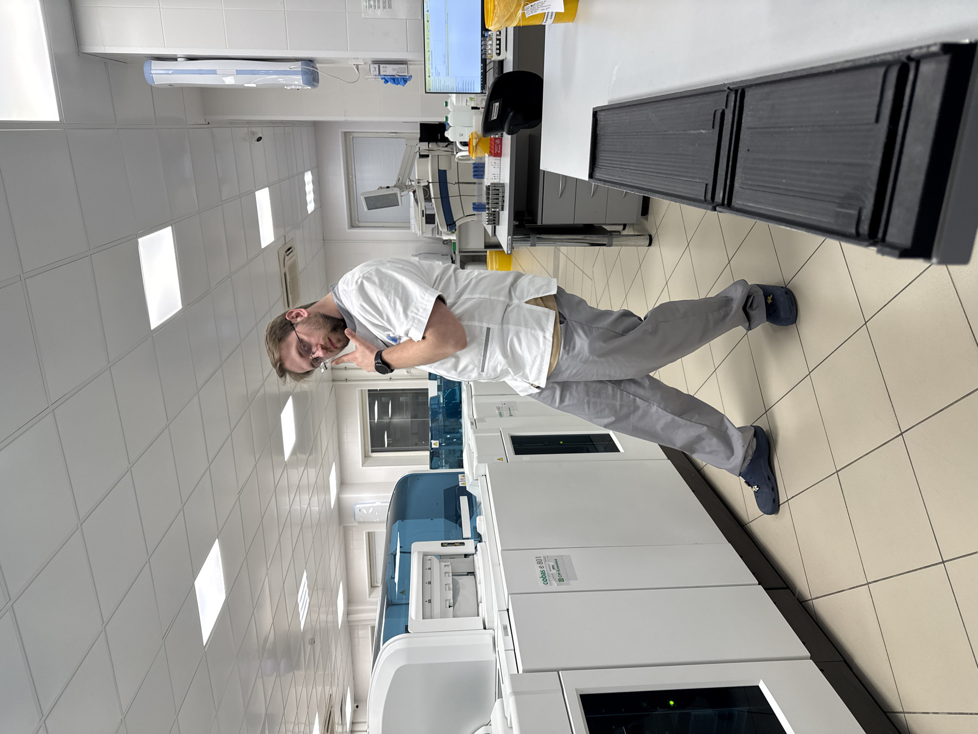 The scene is an indoor laboratory, likely located in Moscow, Russia, although this cannot be definitively confirmed from the image alone. A man, presumably a laboratory worker, is standing in the center of the frame. He is wearing a white lab coat, gray pants, and dark blue shoes, with a wristwatch visible on his left wrist. He has short brown hair and a beard, and is wearing glasses.
Behind him are several large, white medical analyzers, possibly automated blood analyzers or similar diagnostic equipment. These are positioned against a wall with square tiled flooring. The wall is also tiled with white square tiles, and has rows of fluorescent lights in the ceiling. A window is visible in the background.
To the right of the man is a workstation with a computer monitor and various lab equipment. The scene suggests a clinical or research laboratory setting.