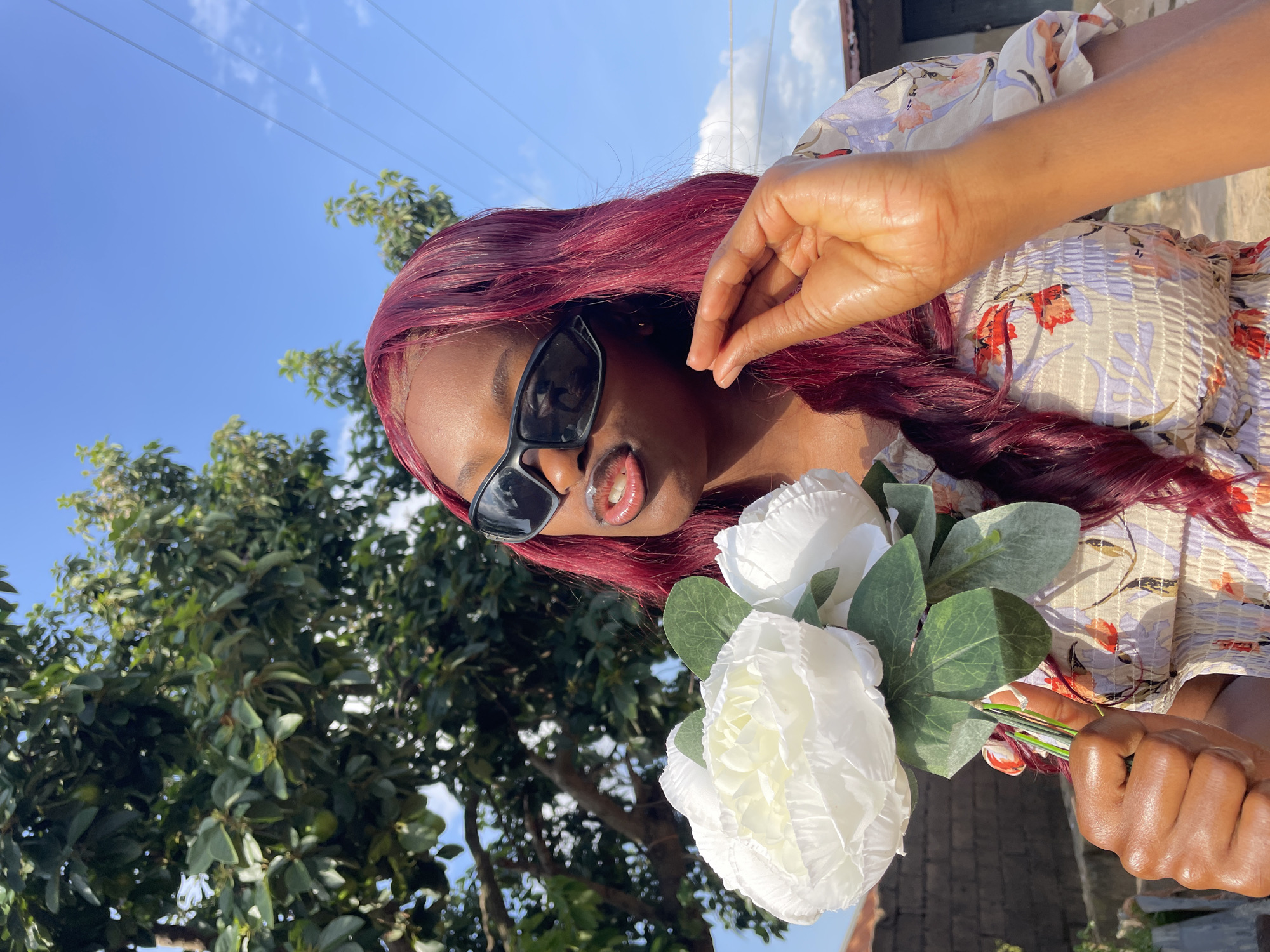 The image features a young Black woman holding a bouquet of white artificial flowers. She is wearing large, dark sunglasses and a floral-patterned top. Her lips are slightly parted, revealing a hint of teeth and possibly lip gloss. Her hair is a vibrant, deep red color and cascades down her shoulders. The image is shot outdoors, with a bright blue sky and some scattered clouds visible in the background. Lush green foliage from trees frames the lower portion of the image, and there are some power lines faintly visible against the sky. The lighting suggests it is daytime, likely sunny. The scene appears to be set in a residential area, possibly a yard or garden. The woman's pose and expression convey a relaxed and perhaps playful mood. The image is oriented vertically, with the sky at the top and the ground and foliage at the bottom. No visible text is present in the image.