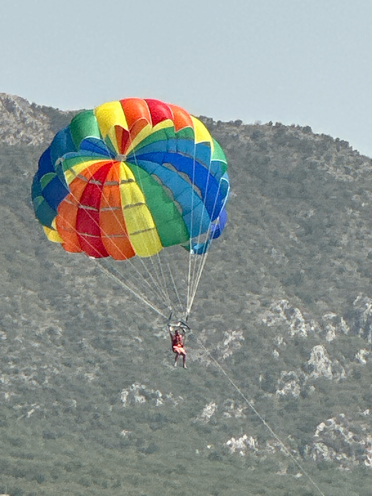 A person is parasailing high above a rocky, vegetated hillside, suspended from a large, colorful parachute. The parachute displays a rainbow pattern of red, orange, yellow, green, and blue segments. The individual is wearing a red garment and is dangling from the parachute's harness. The background consists of a gray sky and a steep, mountainous terrain covered in scrub and rocks. The scene is captured from a distance, emphasizing the vastness of the environment and the height of the parasailer.
