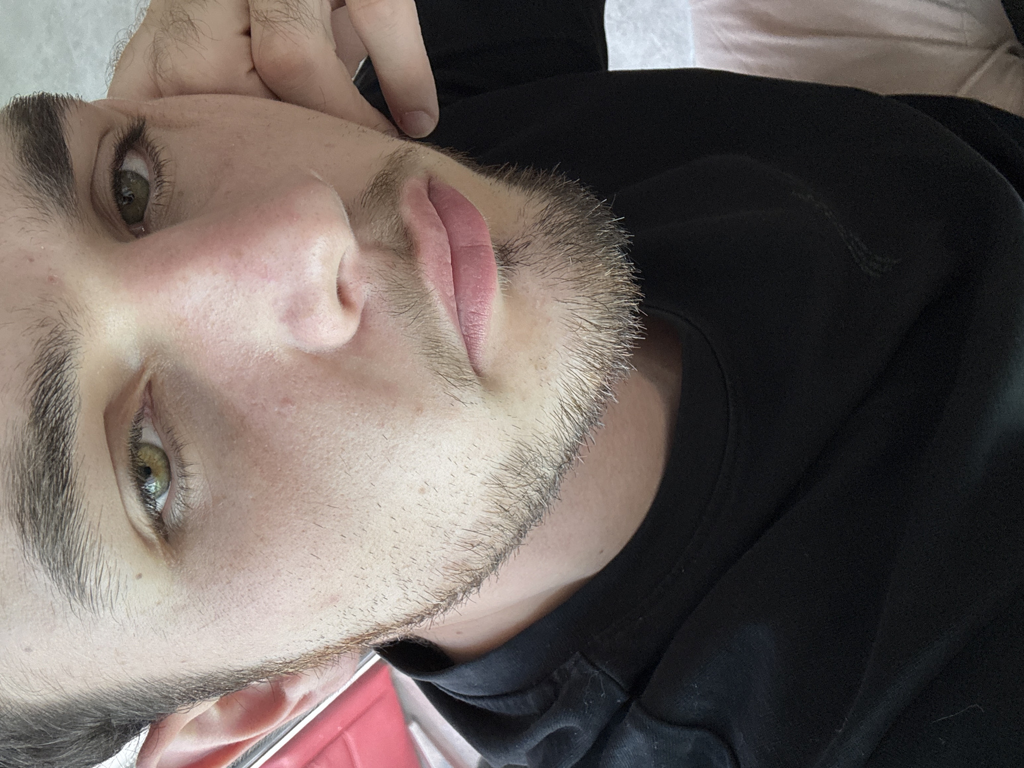 A close-up shot of a fair-skinned man with a sparse beard. He has light brown hair, green eyes, and pink lips. His right hand is near his face. He's wearing a black hooded sweatshirt. The background is a mix of light and dark, with glimpses of a light-colored surface. The scene is unlikely to be in San Bernardino Tlaxcalancingo, Mexico, due to the subject's features, which are not typical of the local population. No specific details or elements indicate the location to be in that particular city in Mexico.