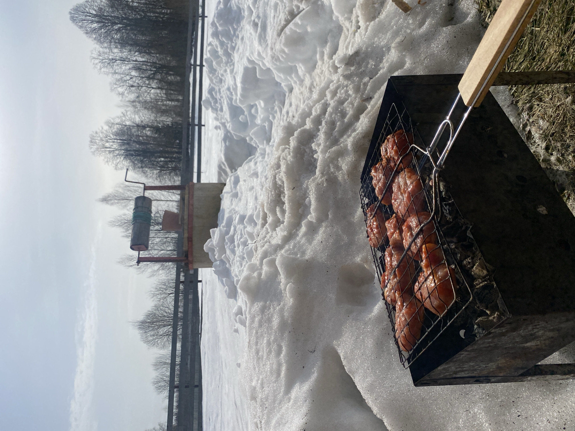 A forensic description of the scene in Zhigalki, Russia, reveals an outdoor cooking setup amidst a snowy landscape under a bright, overcast sky. In the immediate foreground, a black, rectangular metal charcoal grill, or brazier, sits on the ground. A metal grilling basket, with a light-colored wooden handle extending to the right, rests atop the grill, containing multiple pieces of marinated red meat, likely pork or chicken, actively cooking over glowing charcoal. The ground surrounding the grill is a mix of melting snow, some appearing dirty or slushy, and patches of dried brown grass where the snow has receded.
Further into the mid-ground, substantial piles of snow, some glistening, others showing signs of melting and dirt, dominate the terrain. To the right of the frame's center, a traditional water well is visible, featuring a circular concrete base, a red metal A-frame structure, and a wooden drum with a green rope wound around it. A light-brown wooden bucket hangs from the well's mechanism. Beyond the well and snow, a dark, horizontal fence line with visible posts stretches across the middle ground. The background is composed of a dense line of bare deciduous trees, characteristic of a winter or early spring environment. The sky above is bright and largely featureless, suggesting either heavy overcast conditions or a hazy, bright day. No individuals are visible in the scene.